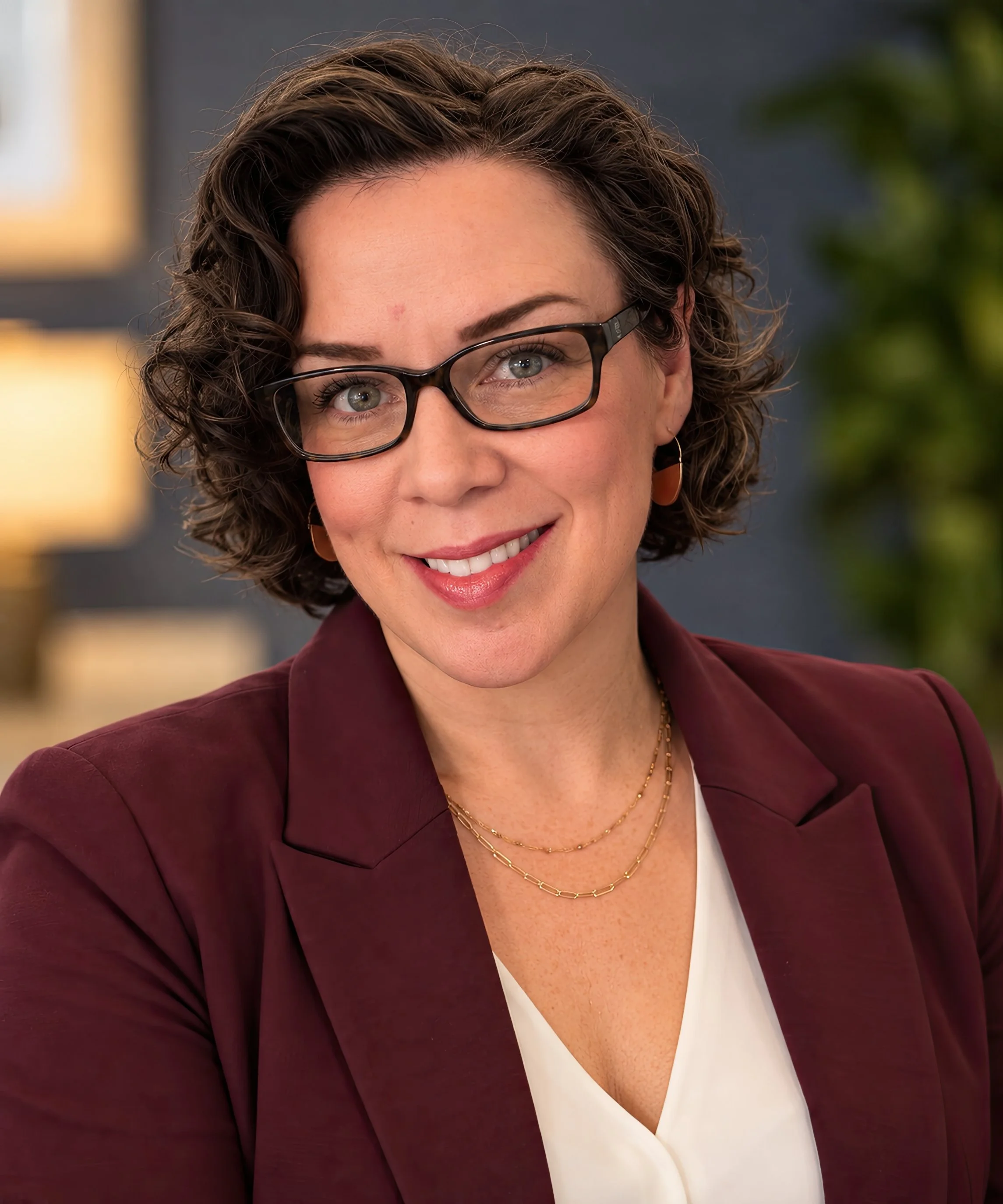 Close-up of a smiling woman with curly brown hair, wearing glasses, gold jewelry, a white top, and a maroon blazer, with a blurred background.