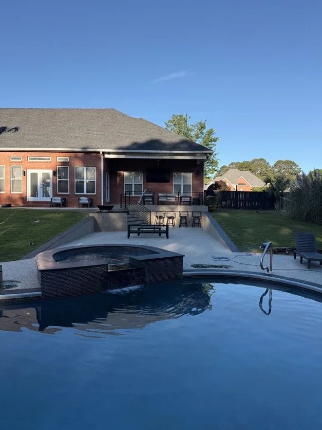 Backyard with in-ground pool and hot tub, resin-bound pebble pool deck and patio designed and installed by Pebble & Resin Co with chairs and a bench, brick house with sliding glass doors, green lawn, and a wooden fence, under a clear blue sky.