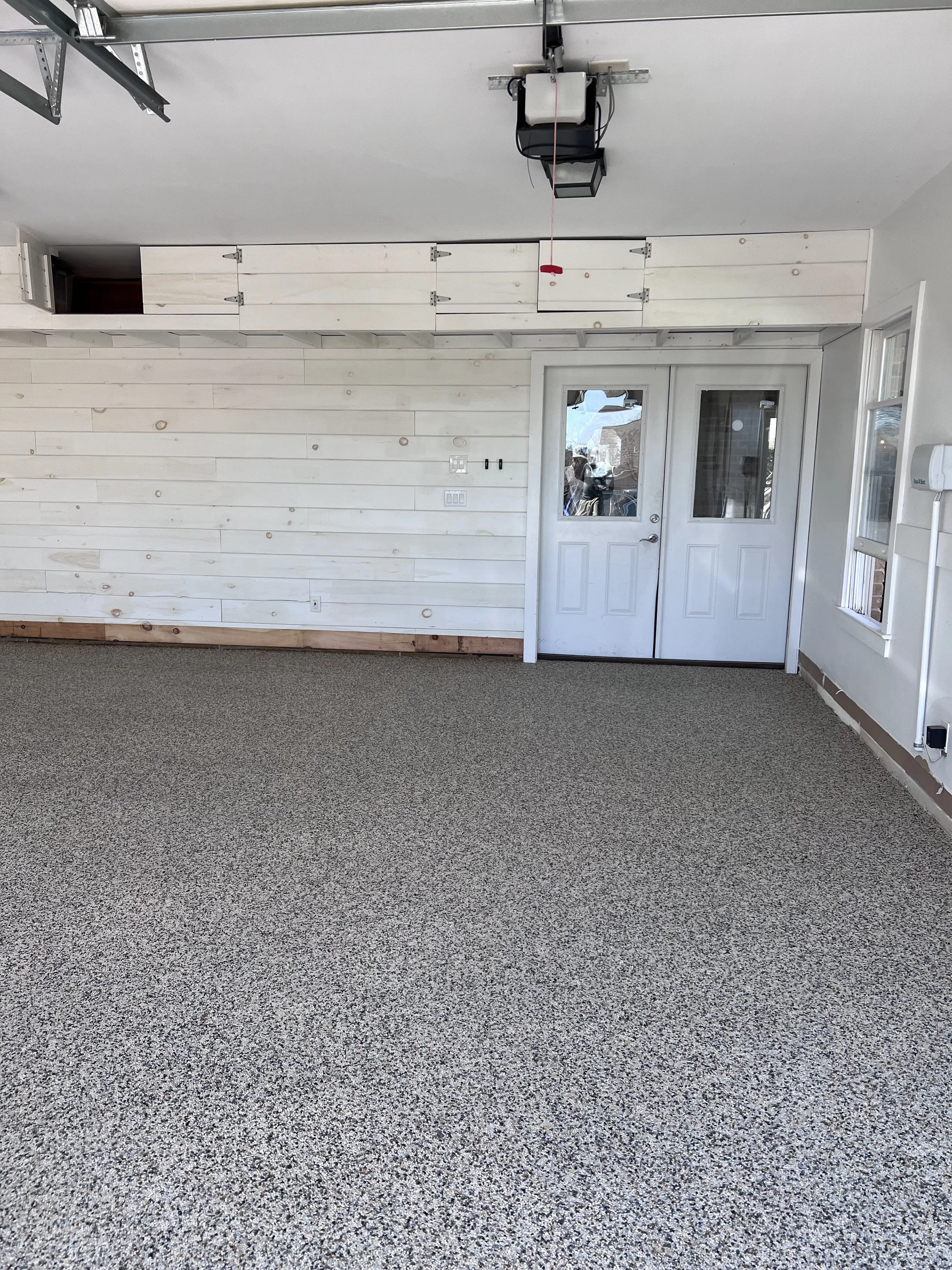 Empty garage with resin-bound pebble flooring designed and installed by Pebble & Resin Co white shiplap wall, and white door with glass window, situated in a residential space.