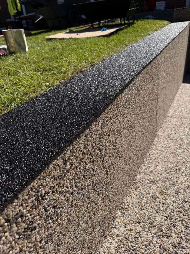 Close-up of a resin-bound pebble pool deck designed and installed by Pebble & Resin Co border with a rough texture, topped with a smooth, black finishing edge, next to a grassy area with gardening tools and a small sandbox.