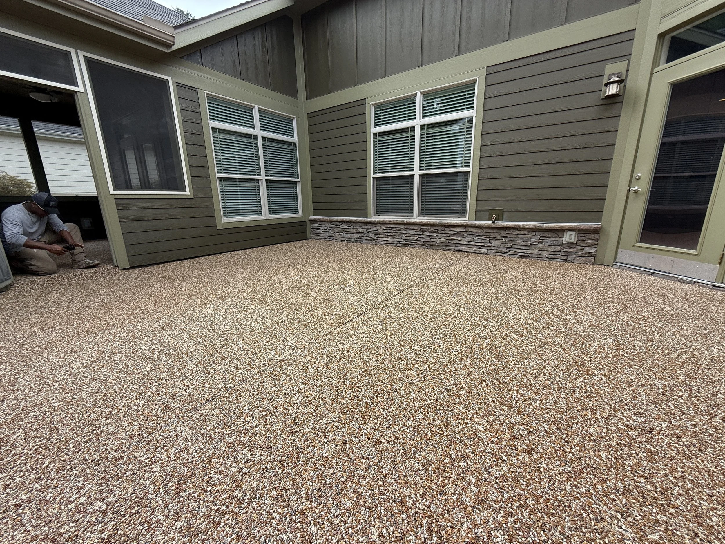 Backyard patio with textured concrete floor, beige siding, multiple windows with blinds, and a small stone wall.