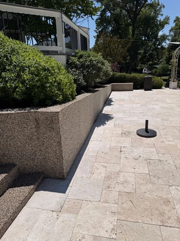 Empty outdoor patio with beige stone pavers, a raised garden bed with green bushes, and a black disc-shaped object on the ground, Epoxy bound pebble stairs designed and installed by Pebble & Resin Co.