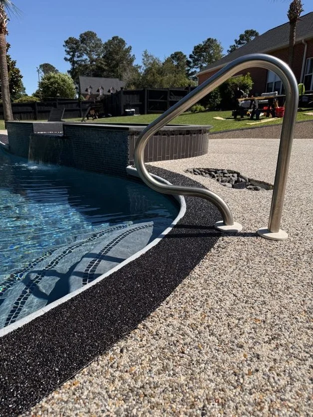 Close-up of an outdoor swimming pool with metal handrails, a black textured pool edge, and a concrete deck, with houses and trees in the background.
