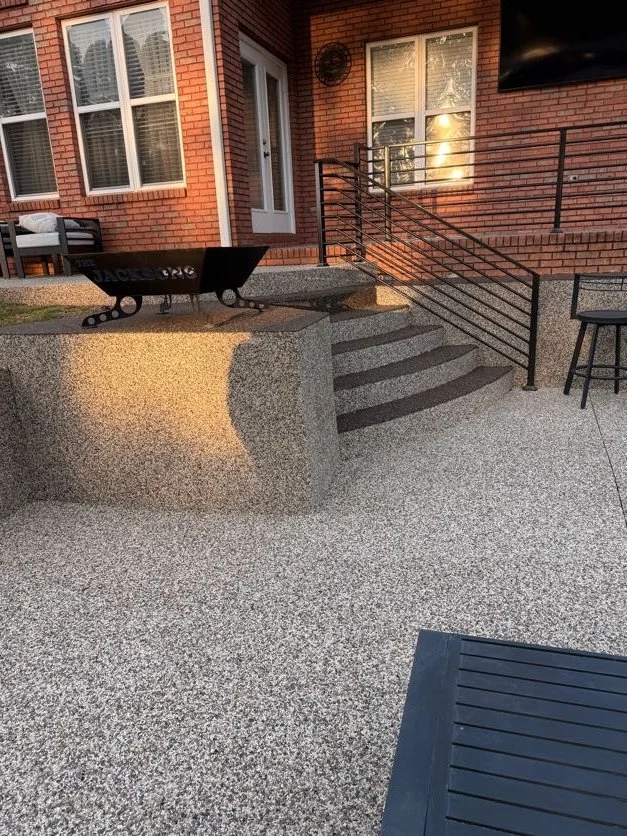 Back patio area with resin-bound pebble flooring and stairs designed and installed by Pebble & Resin Co, brick house, a black metal staircase with handrails leading to a door, a fire pit, and outdoor seating including a black chair and bench,