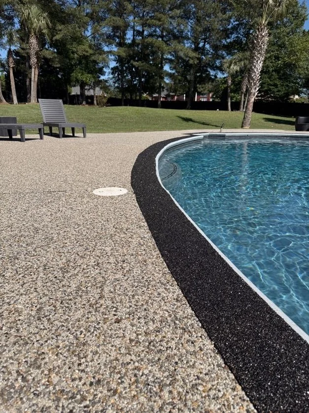 View of a swimming pool with a resin-bound pebble pool deck designed and installed by Pebble & Resin Co, two lounge chairs, grassy area, and trees in the background on a sunny day.