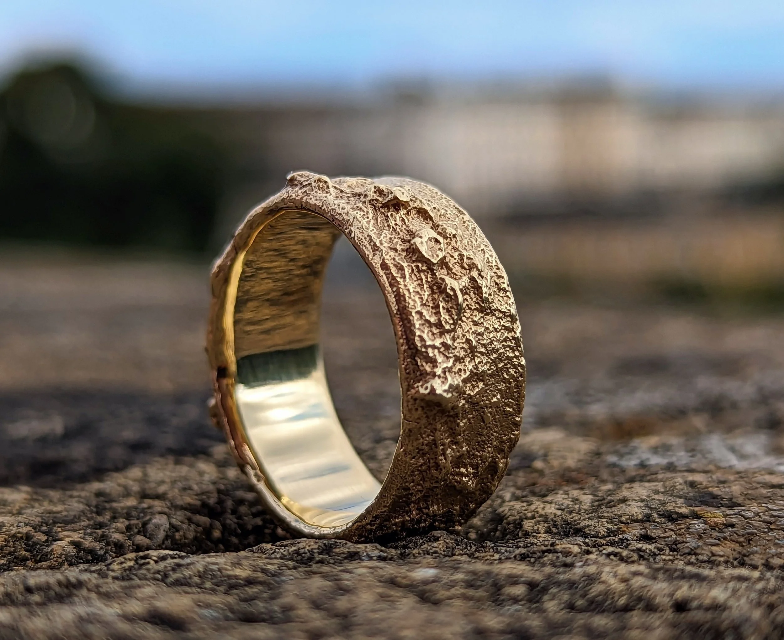 Rockfall range - Close-up of a gold wedding band with a textured surface, resting on a rough, dark surface outdoors with a blurred background.