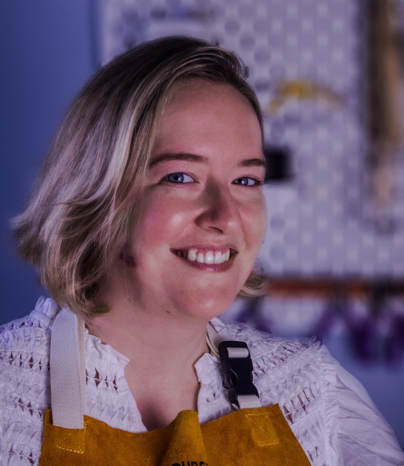 A woman with short blonde hair, smiling, wearing a white textured blouse and a yellow apron, in what appears to be a kitchen or bar setting.