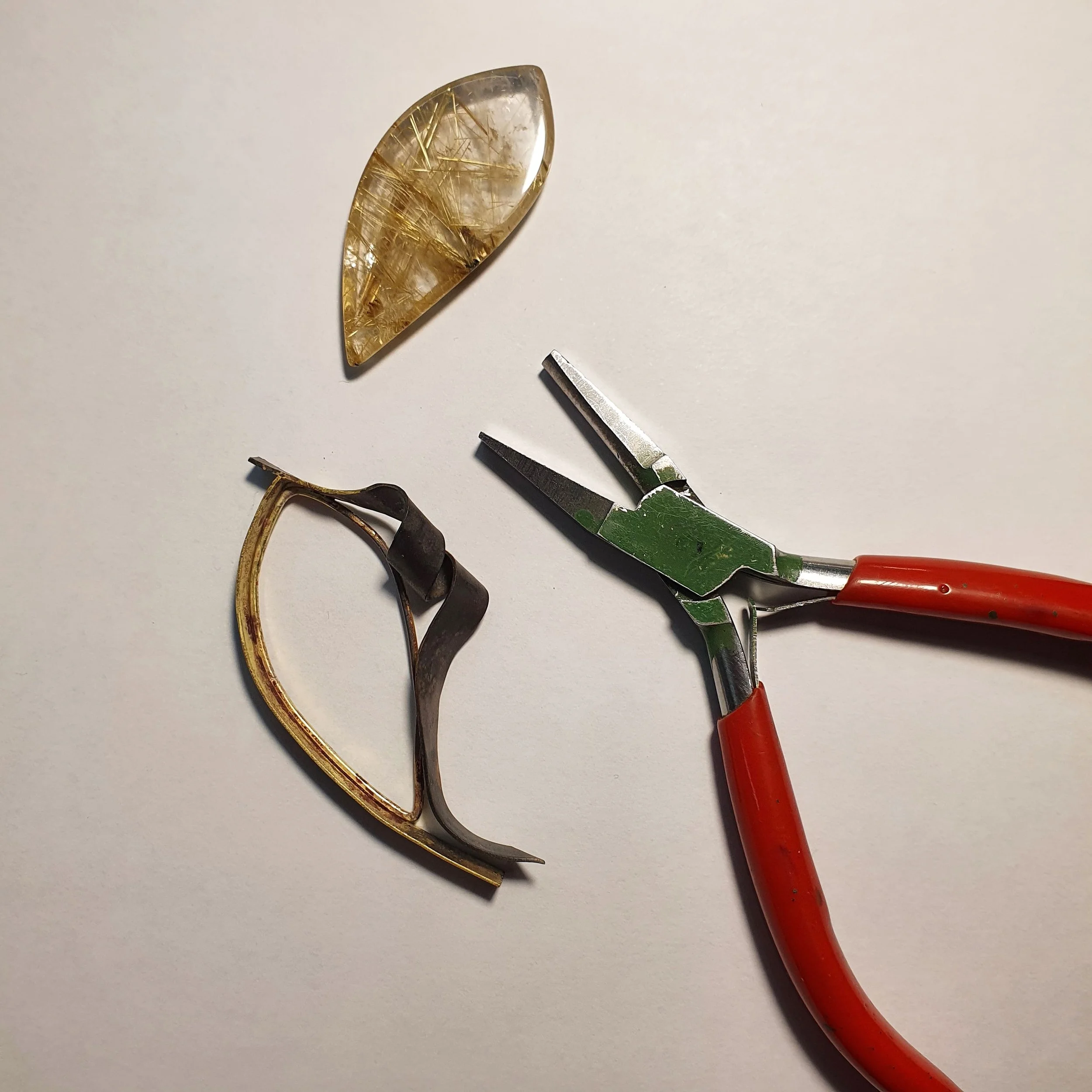 A close-up view of a small fossilized fish preserved inside a translucent, elongated gemstone, with a pair of small pliers nearby on a white surface.