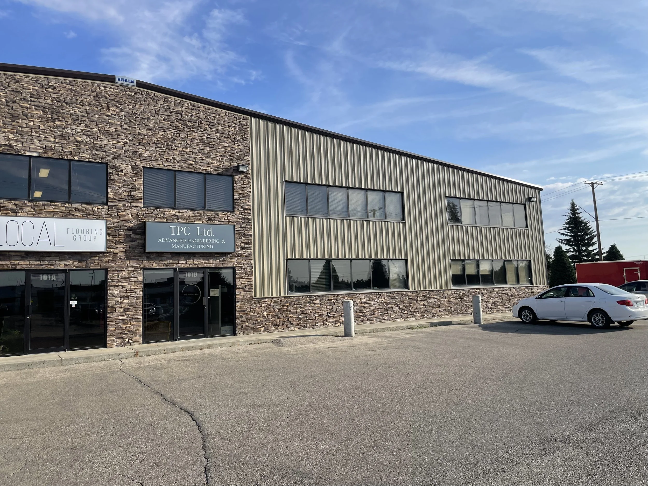 An industrial building with a stone and metal exterior, several windows, and a parking lot with a white car, under a blue sky with clouds.