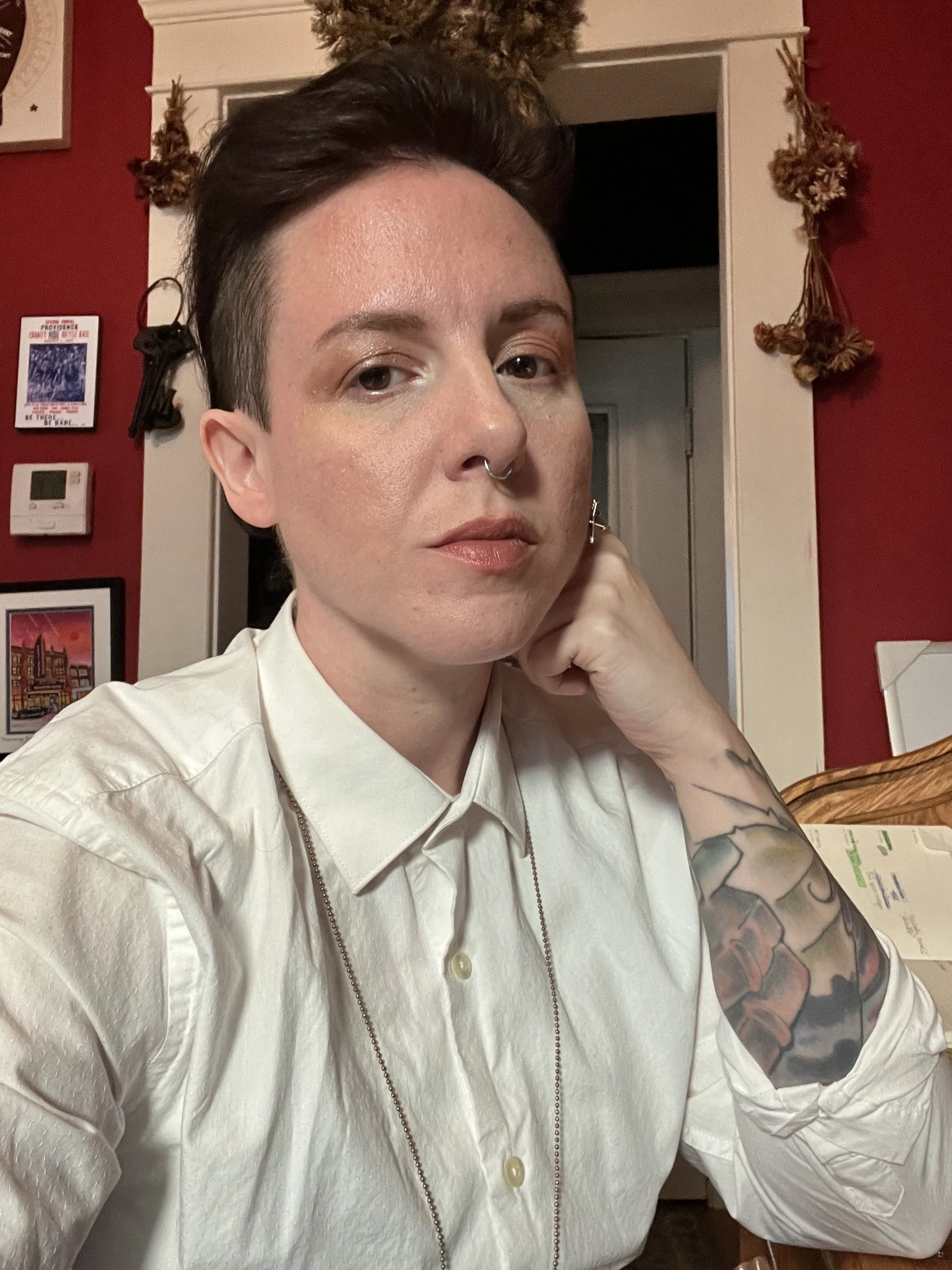 Headshot of Erin Vachon wearing a white shirt with tattoos on their arm, sitting at a table in a decorated room.