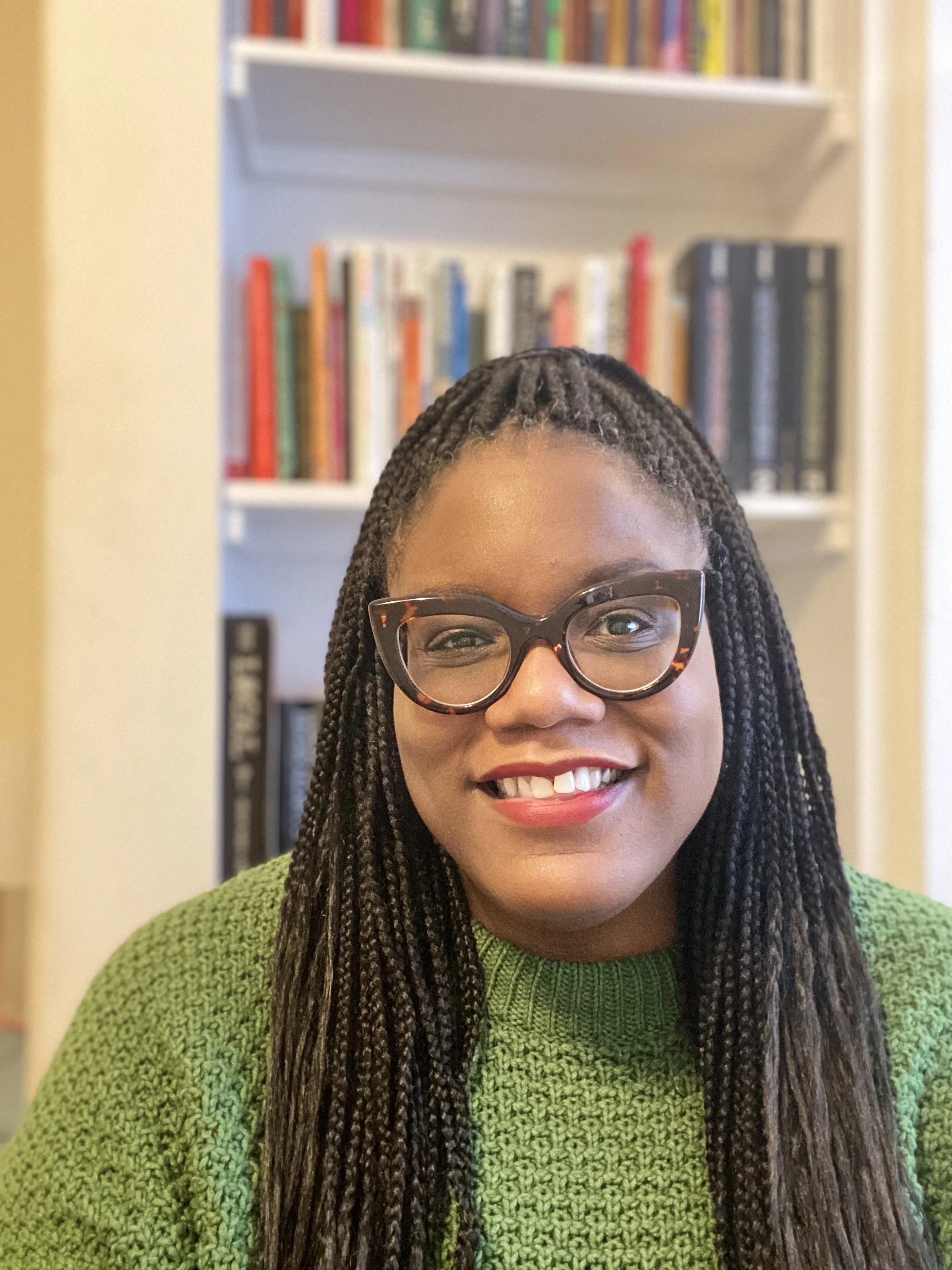Headshot of Athena Dixon wearing glasses and a green sweater, with a bookshelf filled with colorful books in the background.