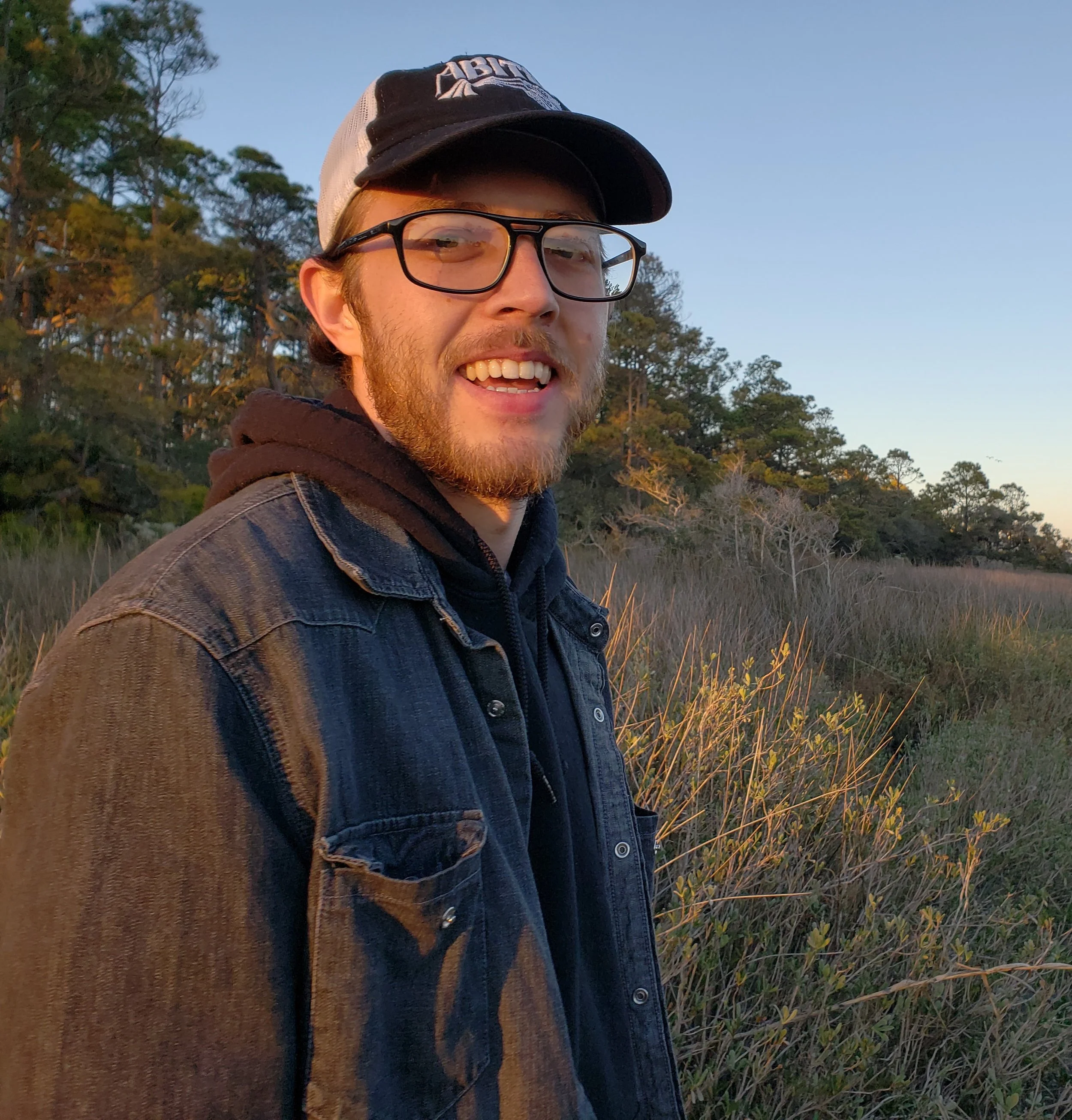 Headshot of Gage Saylor, smiling in a natural outdoor setting during sunset, with trees and grassy fields in the background.