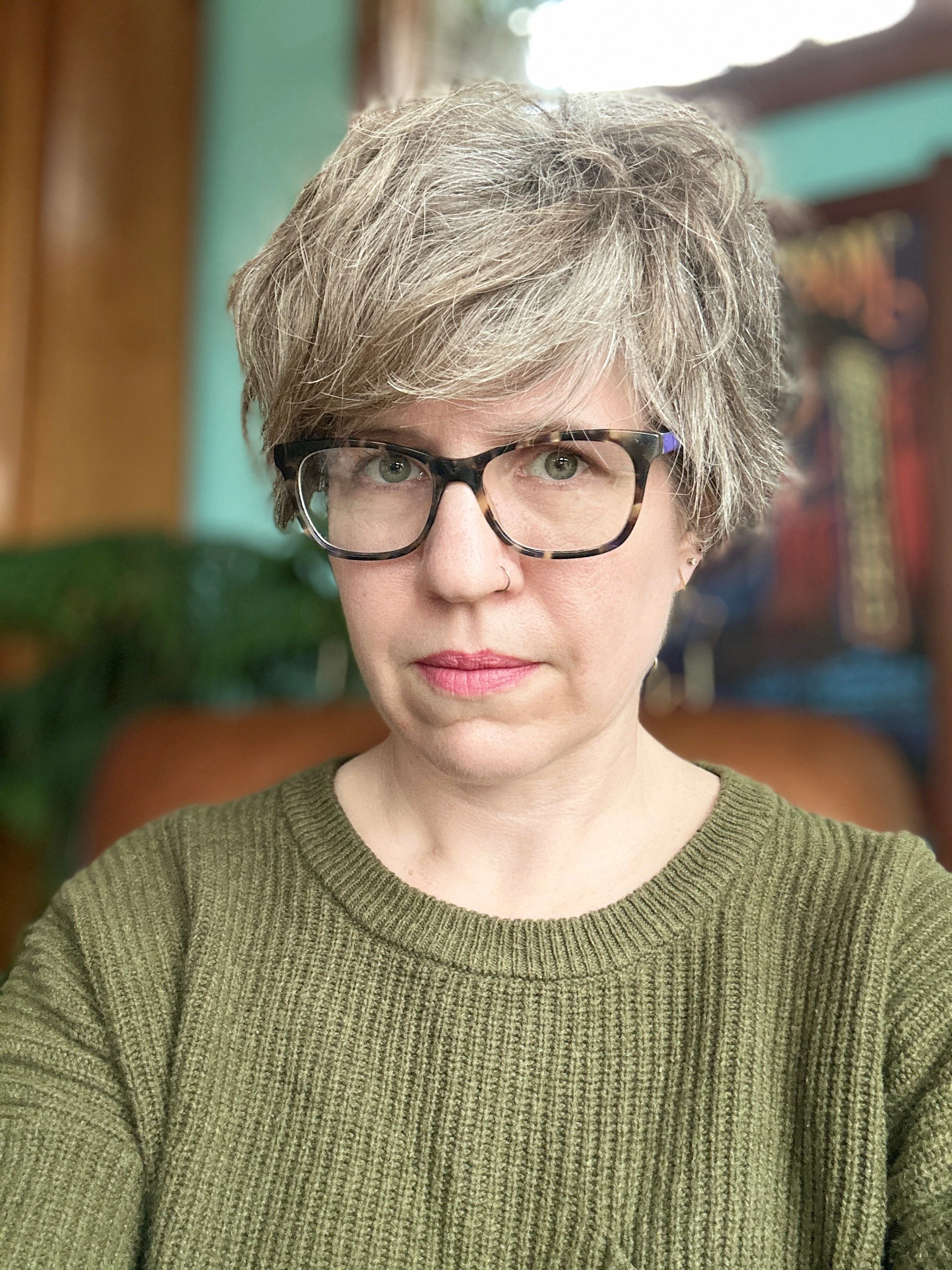 Hearshot of Kristine Langley Mahler. She's wearing glasses with tortoiseshell frames, and wearing an olive-green knitted sweater. Background appears to be indoors with colorful decor.