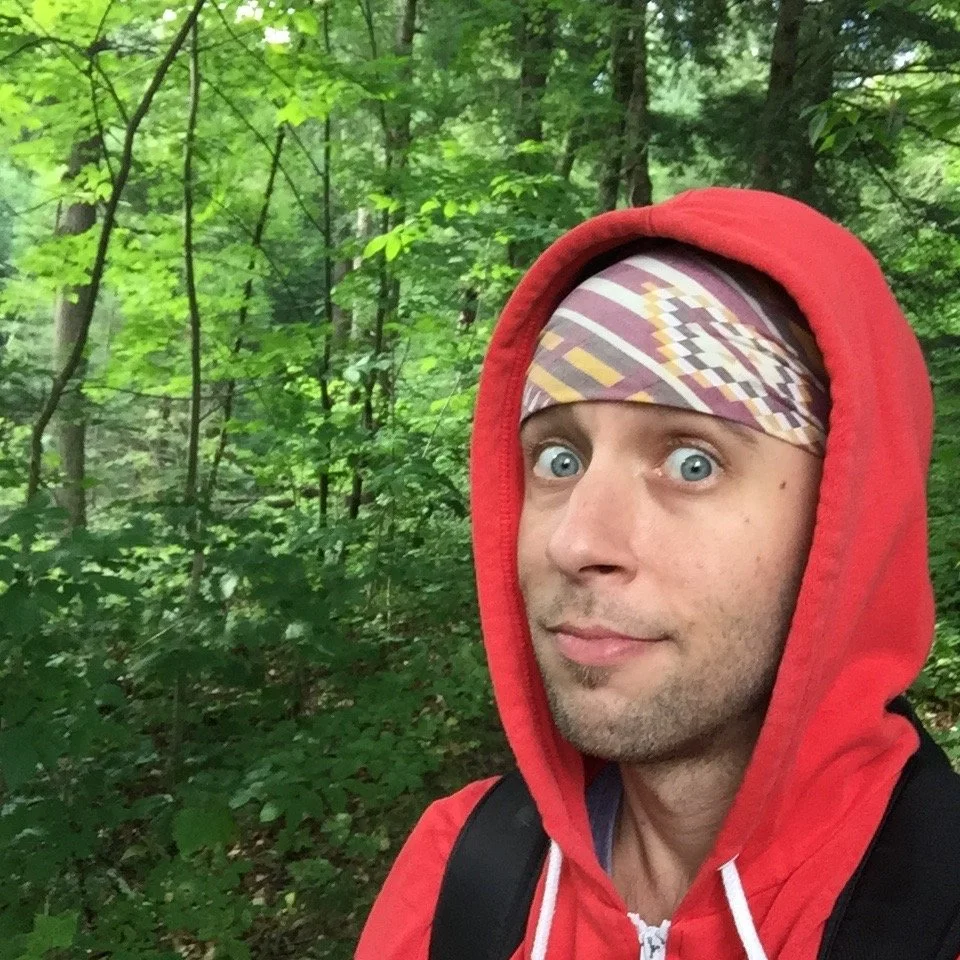 Headshot of David Wojciechowski in a red hoodie and a patterned bandana taking a selfie in a lush green forest.