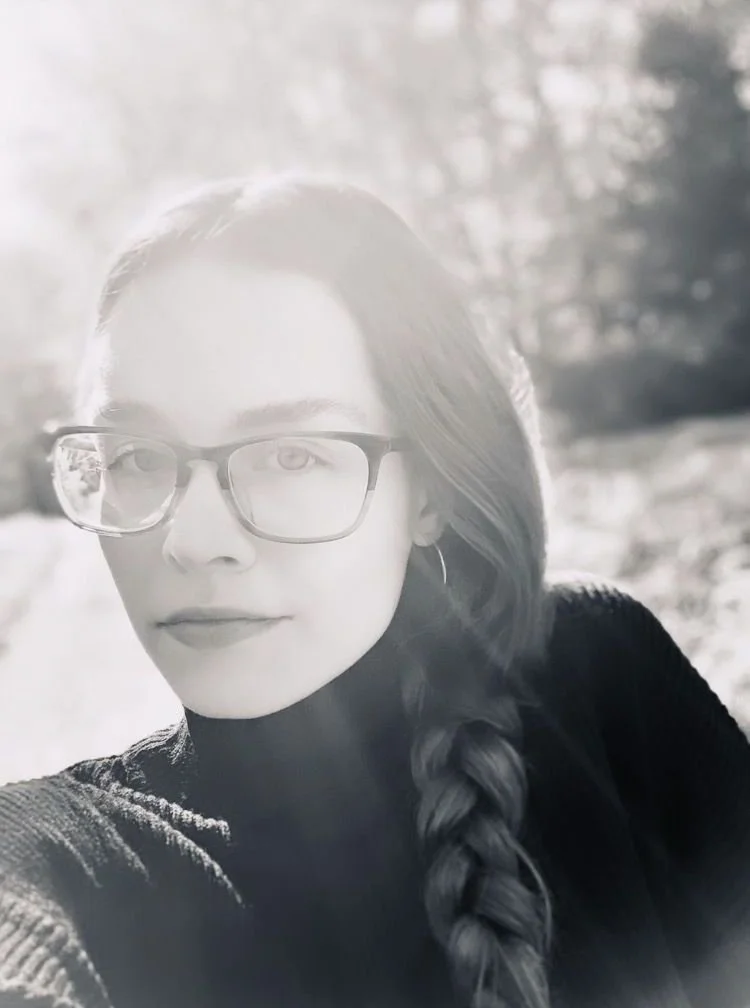 A black-and-white headshot of Abigail Oswald. She's wearing glasses, long hair in a braid, outdoors in a natural setting with sunlight and trees in the background.