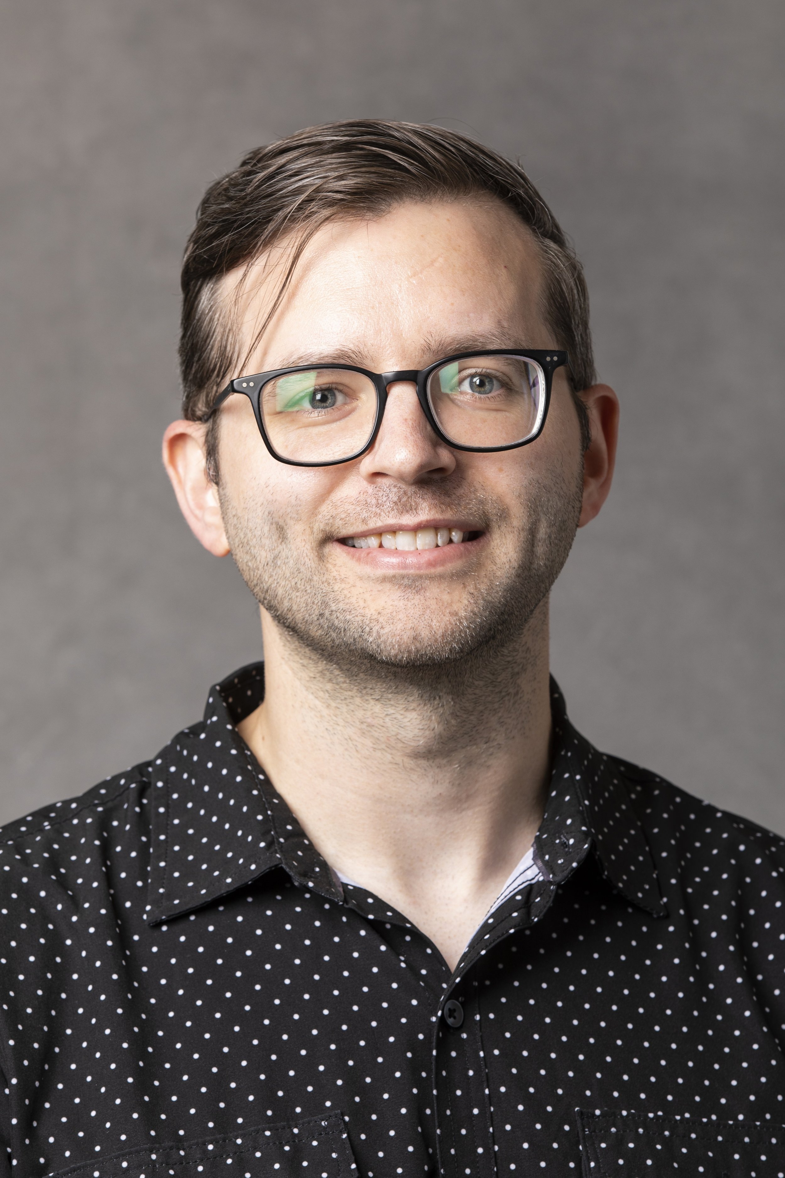 Headshot of Caleb Tankersley wearing a black button-up shirt with white polka dots, against a gray background.