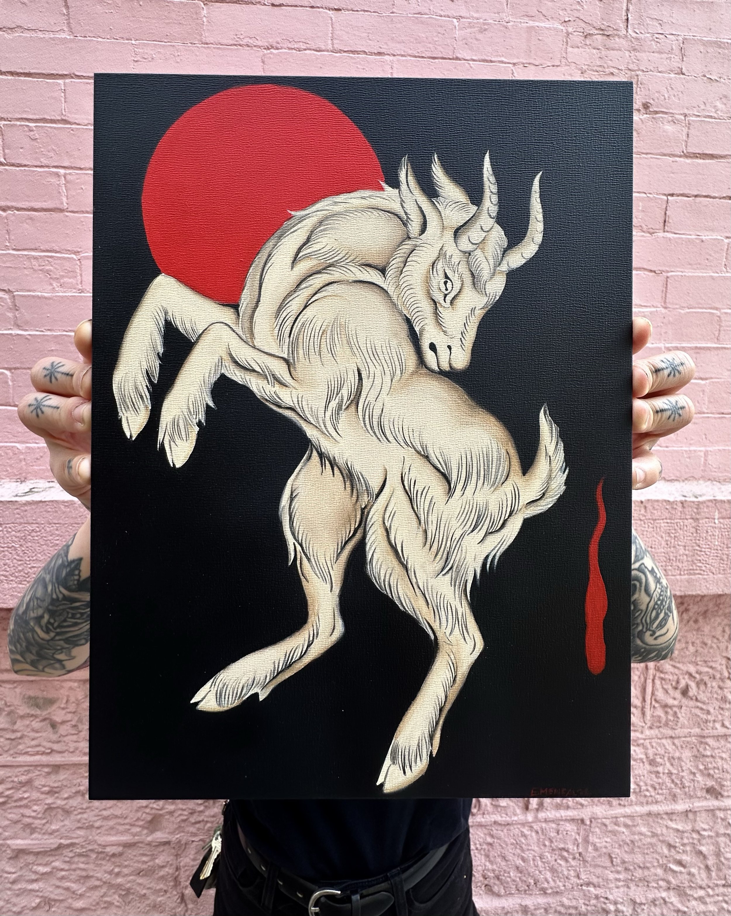 A person holding a black canvas with a white unicorn illustration and a large red circle in the background, against a pink brick wall.