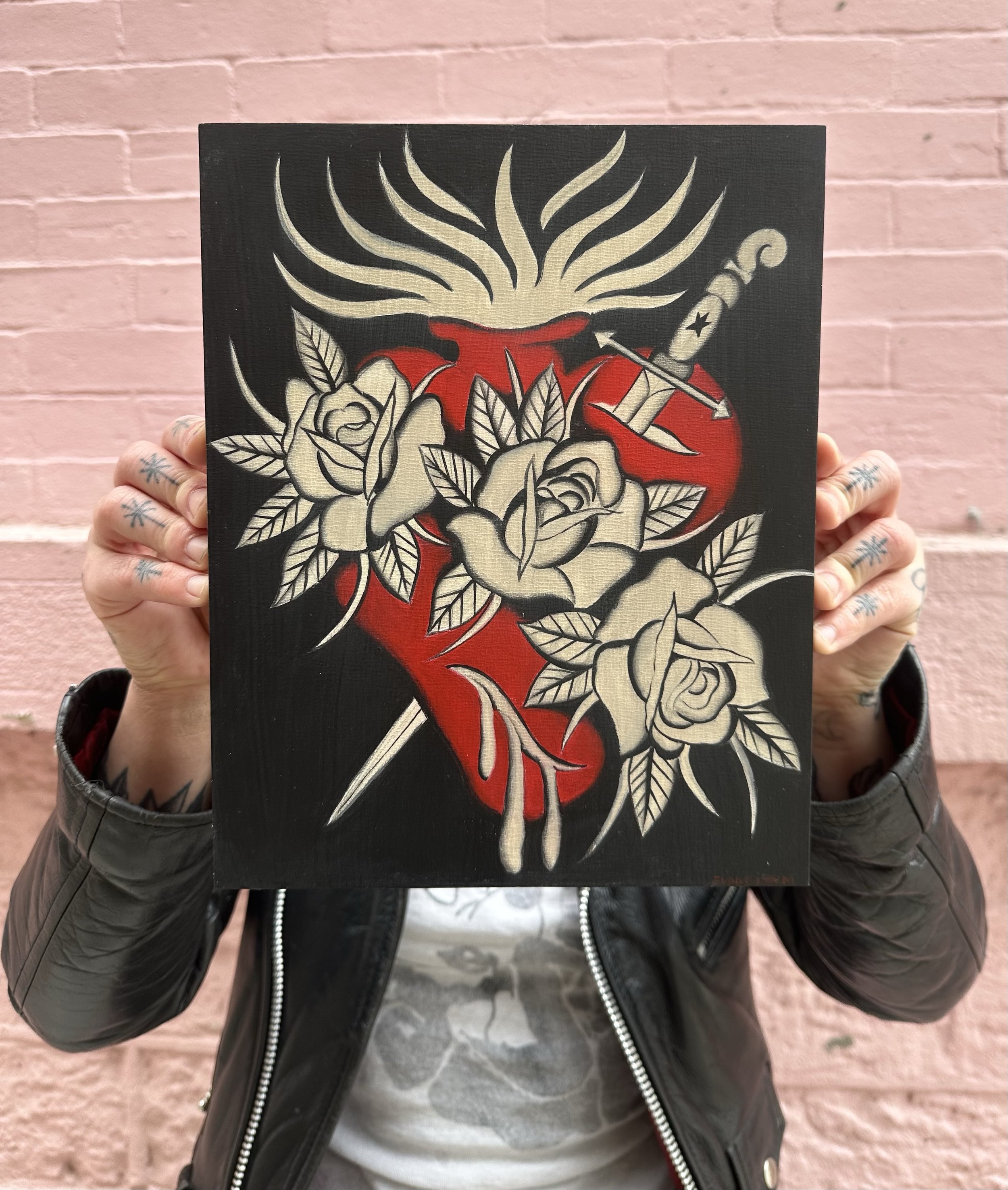 Person holding a framed artwork featuring a heart with roses, a fire torch, and flames, against a pink brick wall background.