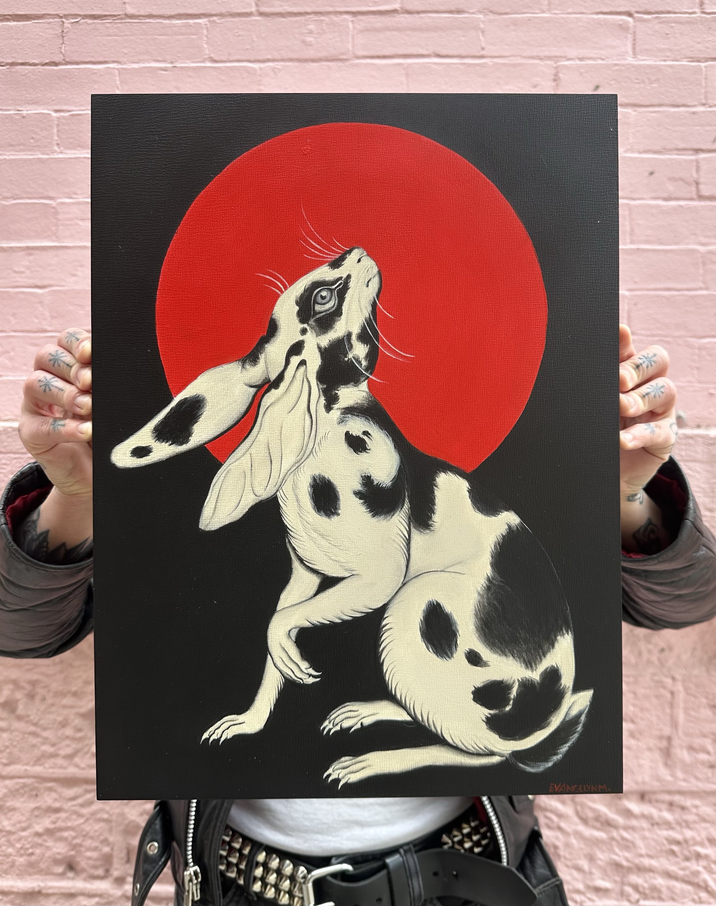 A person holding a painting of a Dalmatian dog with a red circle background in front of a pink brick wall. The person's hands, which have tattoos, are visible on each side of the painting.
