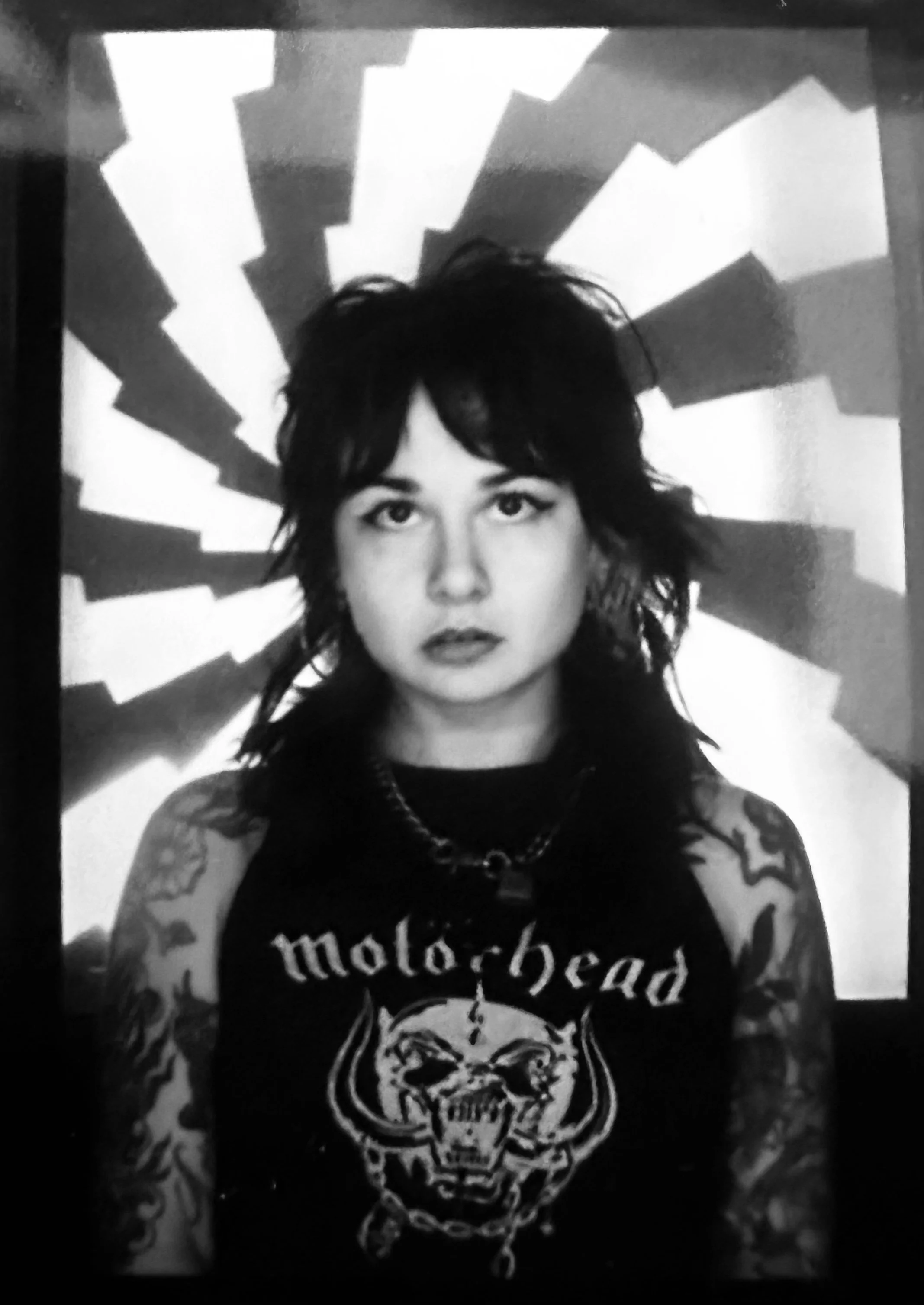 Black and white photo of a woman with wavy dark hair. She has tattoos on her arms and is wearing a sleeveless shirt with a graphic design of a snarling skull and the word 'motorhead.' She has a chain necklace and an intense expression. The background features a radial pattern of black and white shapes.