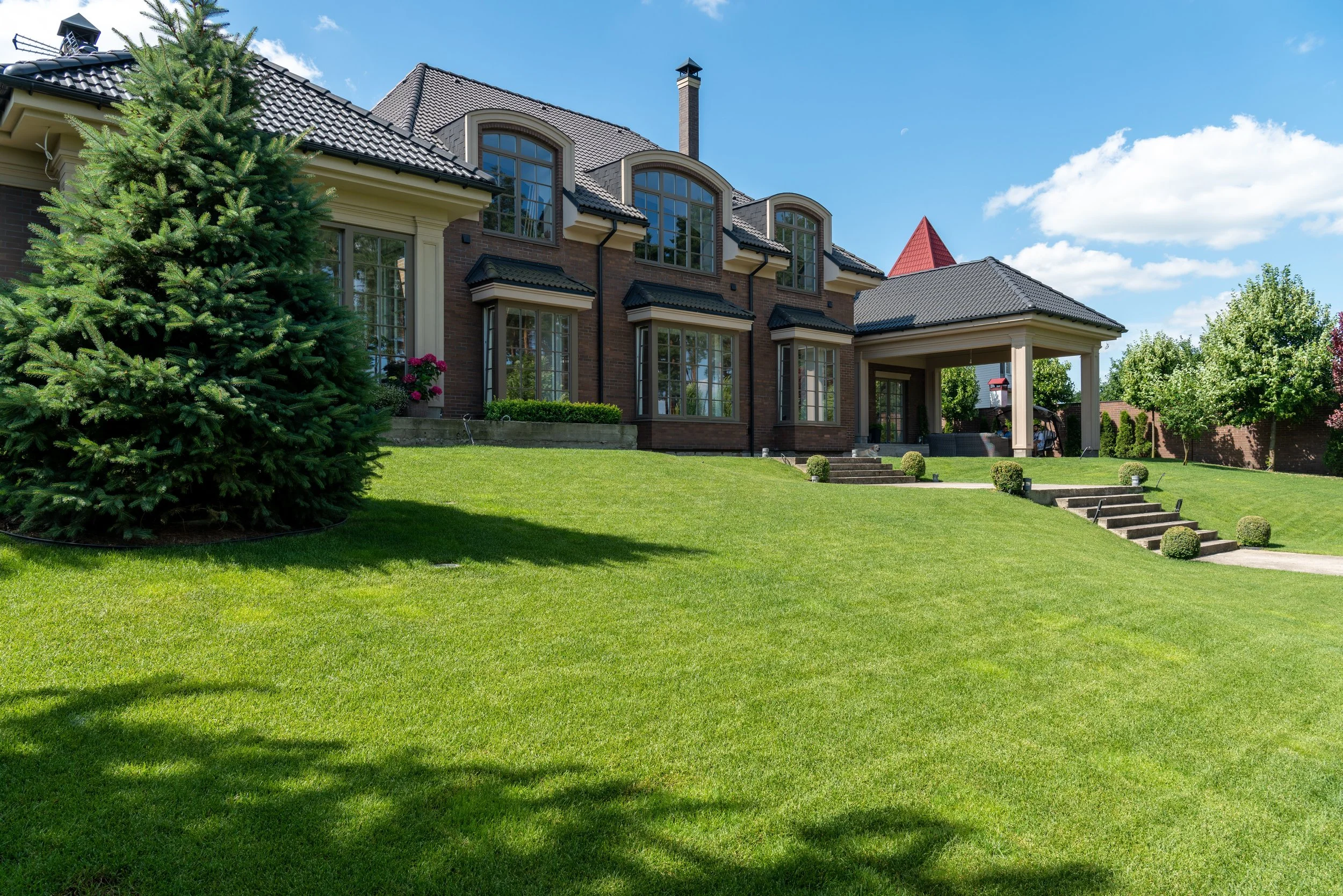 A large brick house with a gray tiled roof and multiple large windows, surrounded by a well-kept green lawn, small trees, and flowering plants, under a bright blue sky.