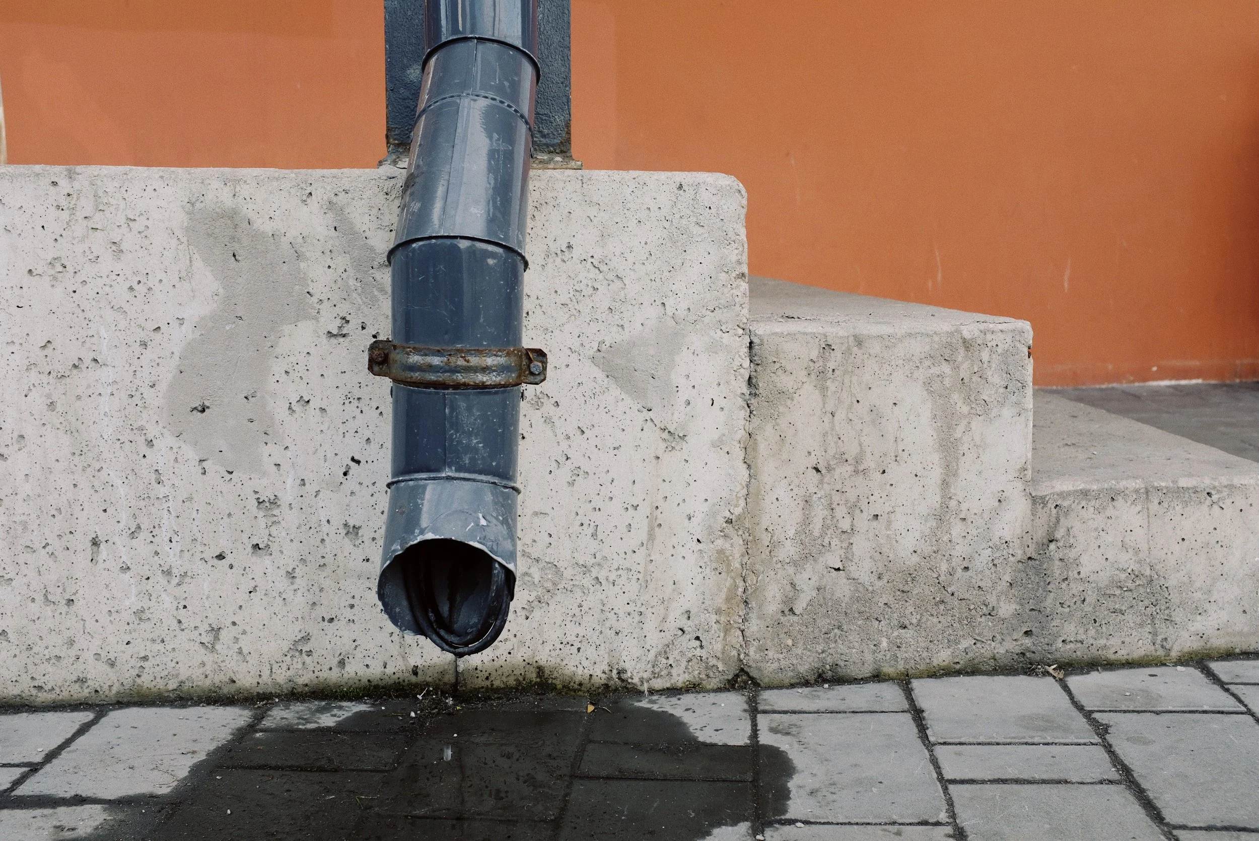 A blue drainpipe attached to a concrete wall, with a rusted metal clamp securing it. The drainpipe extends downward, with an opening at the bottom. The concrete wall shows signs of wear and discoloration, with a background of a reddish-orange wall and tiled pavement below.