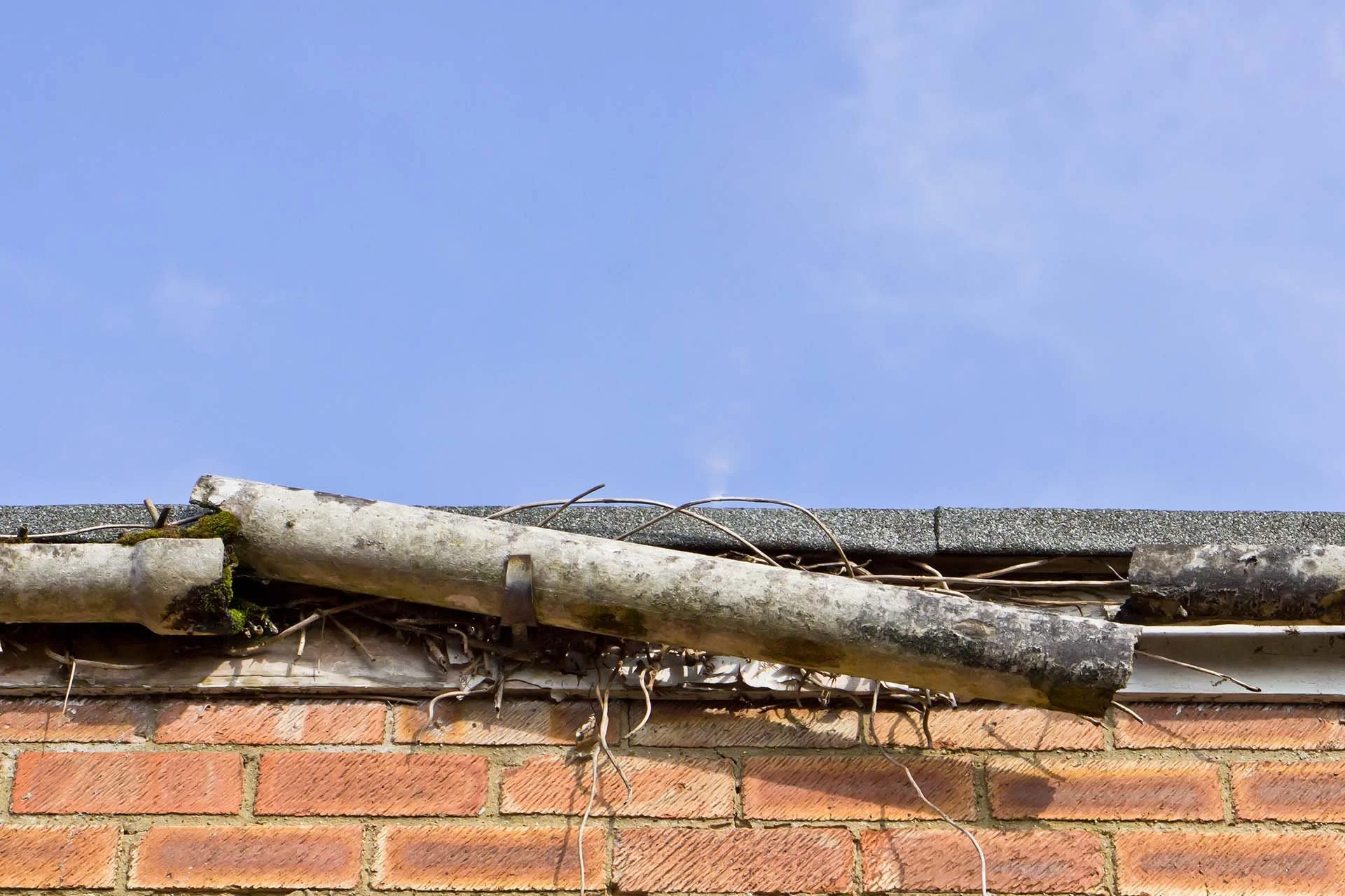 Close-up of a worn roof with old, moss-covered tiles and exposed wires, brick wall underneath, against a blue sky with a few clouds.