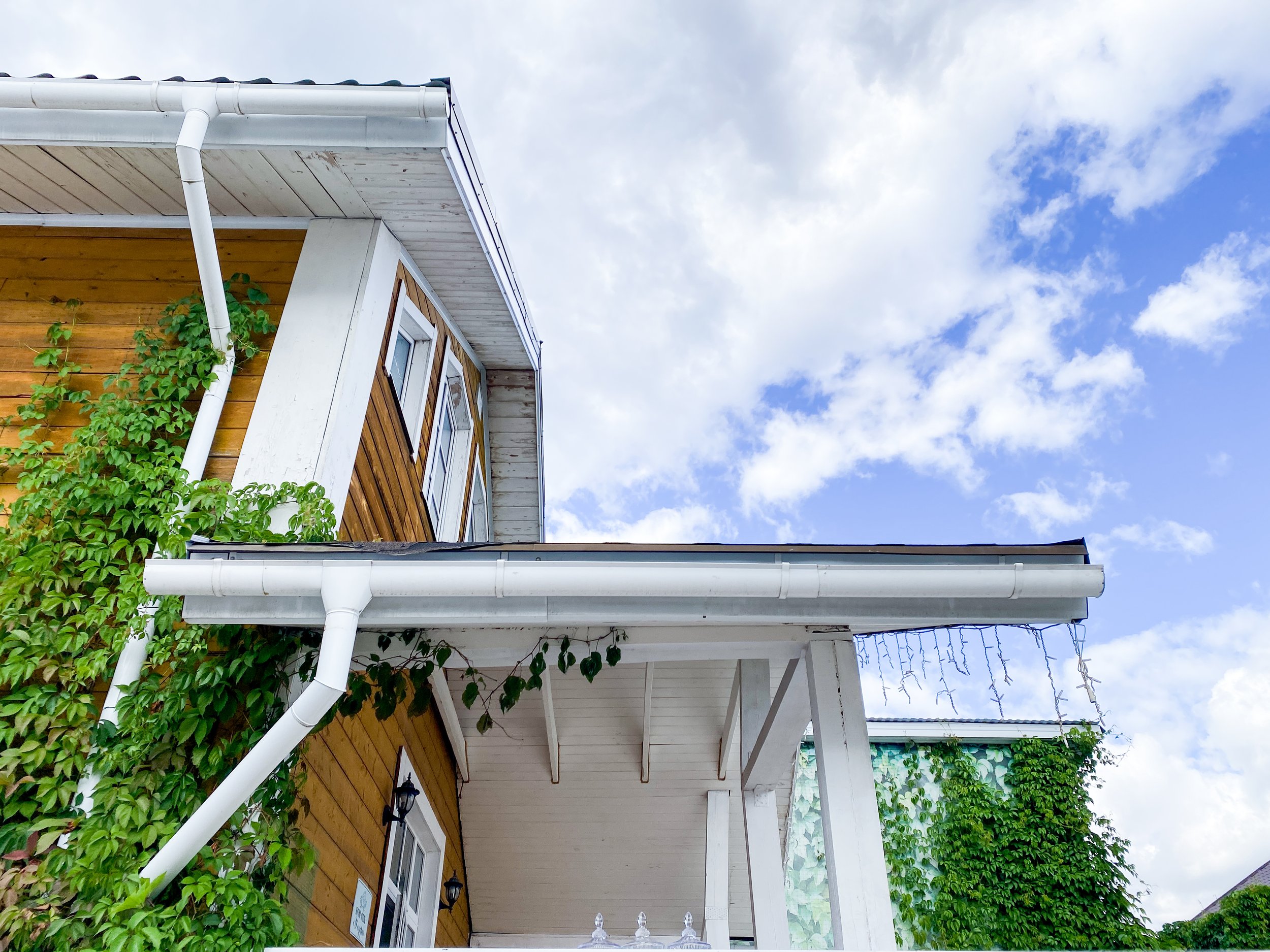 The image shows the upper section of a house with wooden siding, a white gutter and downspout, and a small balcony or porch area with a roof. There are green plants with large leaves growing around the house. The sky in the background is partly cloudy with patches of blue visible.