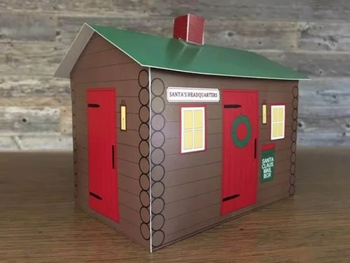 A small cardboard model of Santa's workshop with a brown log cabin design, red doors, yellow windows, a green roof, and signs indicating it is Santa's headquarters and a Santa Claus delivery box.
