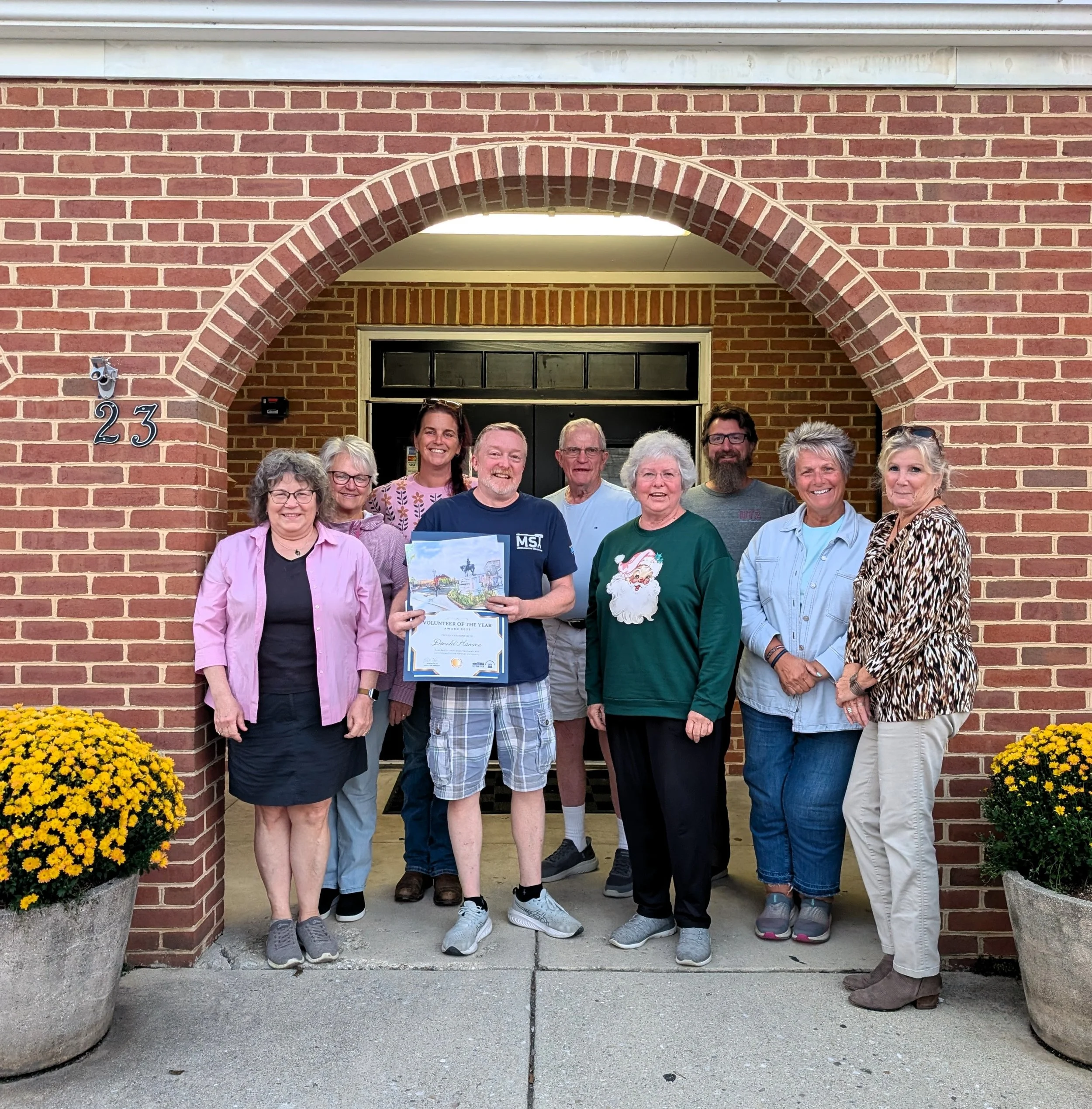 Group of people standing in front of a brick building with an arched entryway, holding a certificate, with yellow mums in pots at the sidewalk corners.