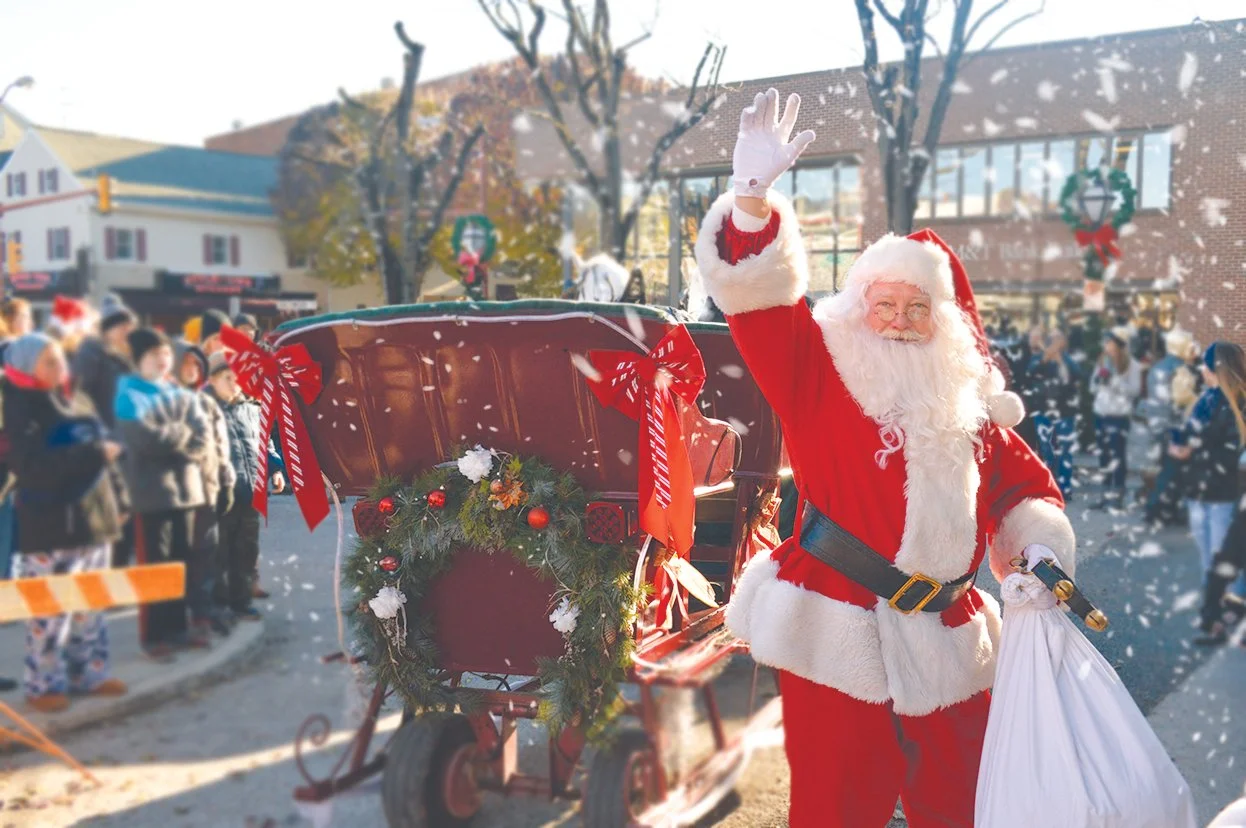 Santa Claus waving at a Christmas parade, standing next to a decorated horse-drawn carriage, with onlookers in winter clothing and Christmas decorations in the background.