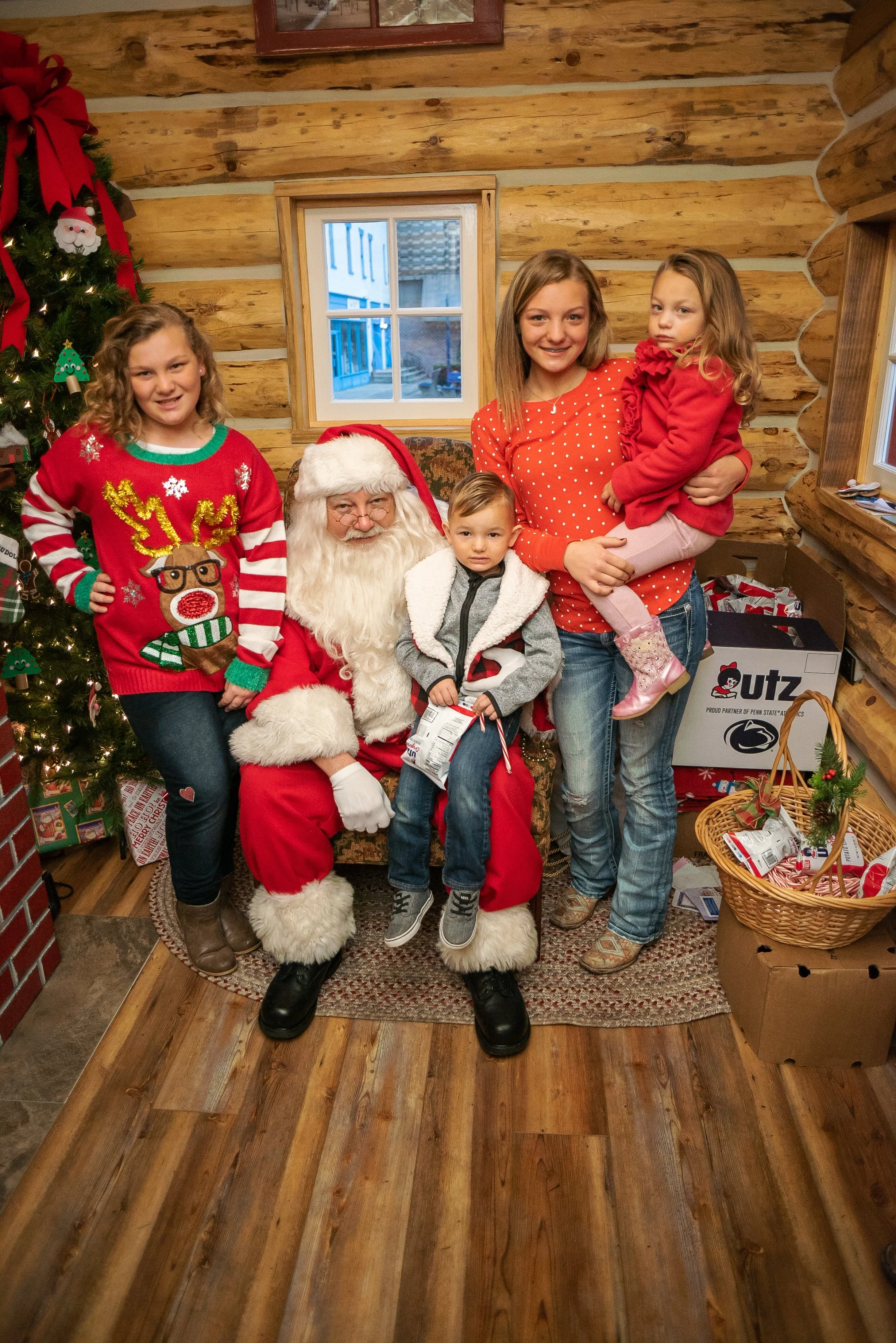 A group of five children with Santa Claus in a cozy, wood-paneled room with Christmas decorations, including a tree and presents.