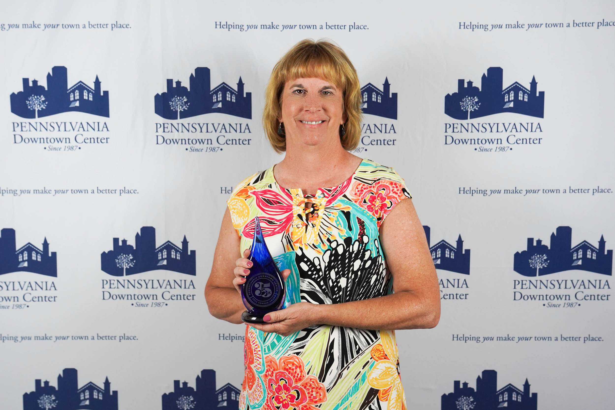 Woman with short blonde hair smiling, holding an award, dressed in a multicolored floral dress, standing in front of a backdrop with Pennsylvania Downtown Center logos.