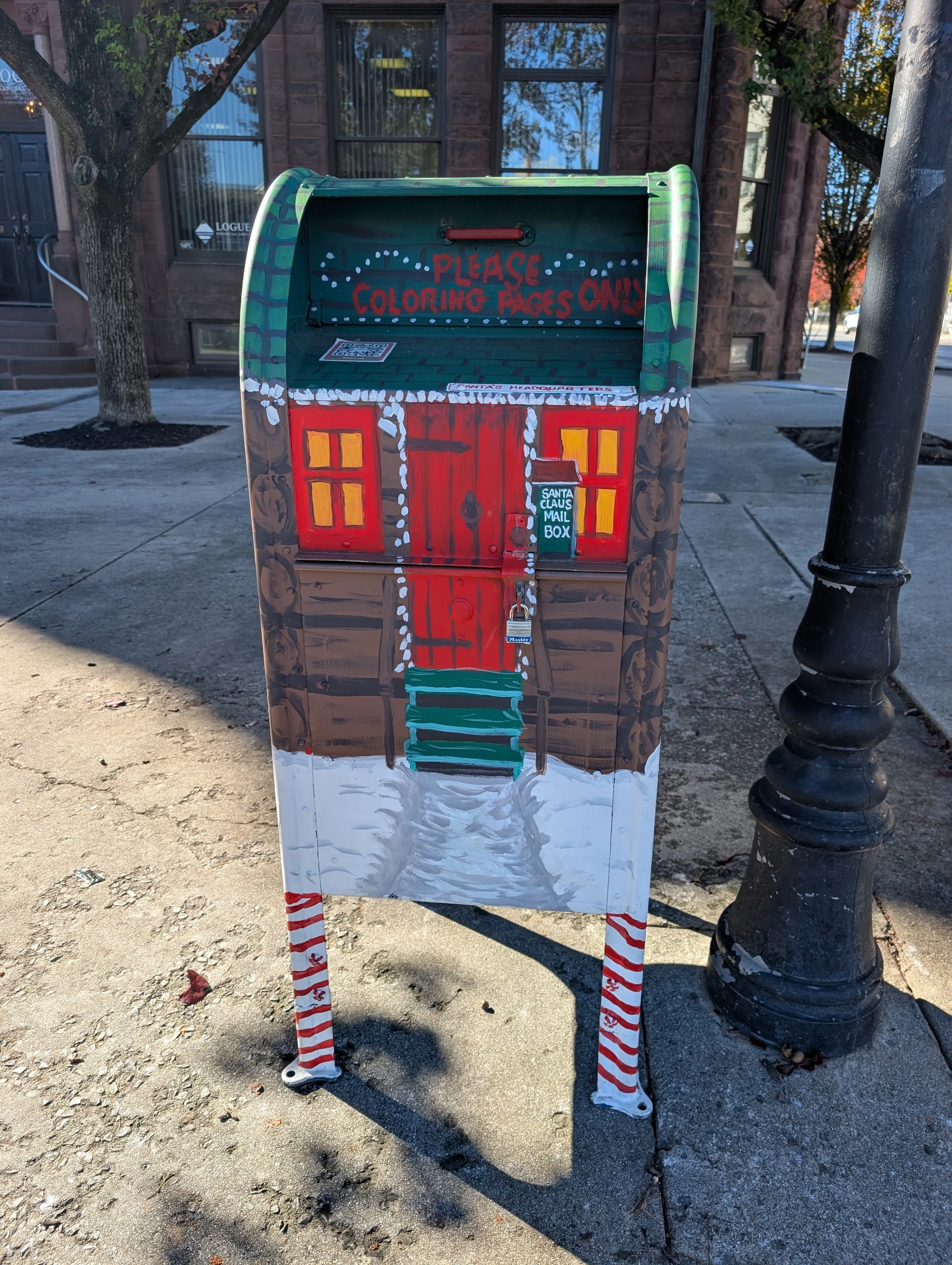 A decorated mailbox resembling a gingerbread house with a red door, yellow window panes, white icing accents, and a 'Santa Claus Mail Box' sign. The mailbox is outdoors on a sidewalk, with a tree and a stone building in the background.
