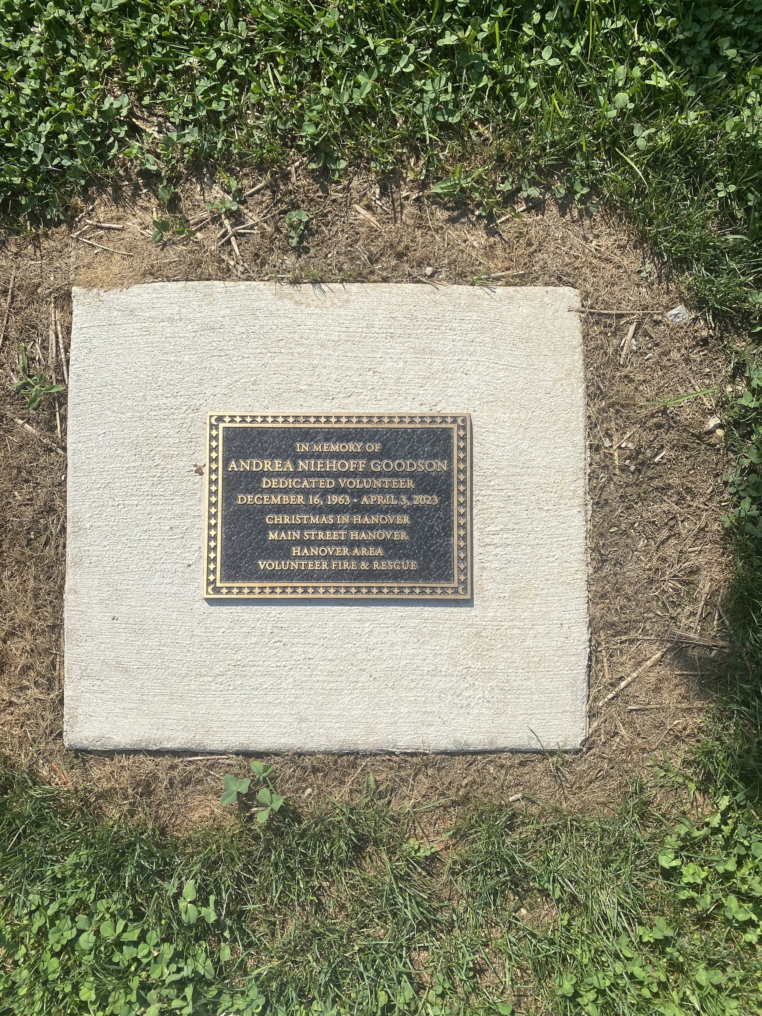 Memorial plaque dedicated to Andrea Niehoff Goodson, a volunteer who passed away on April 3, 2023, located on a concrete base surrounded by grass and small plants.