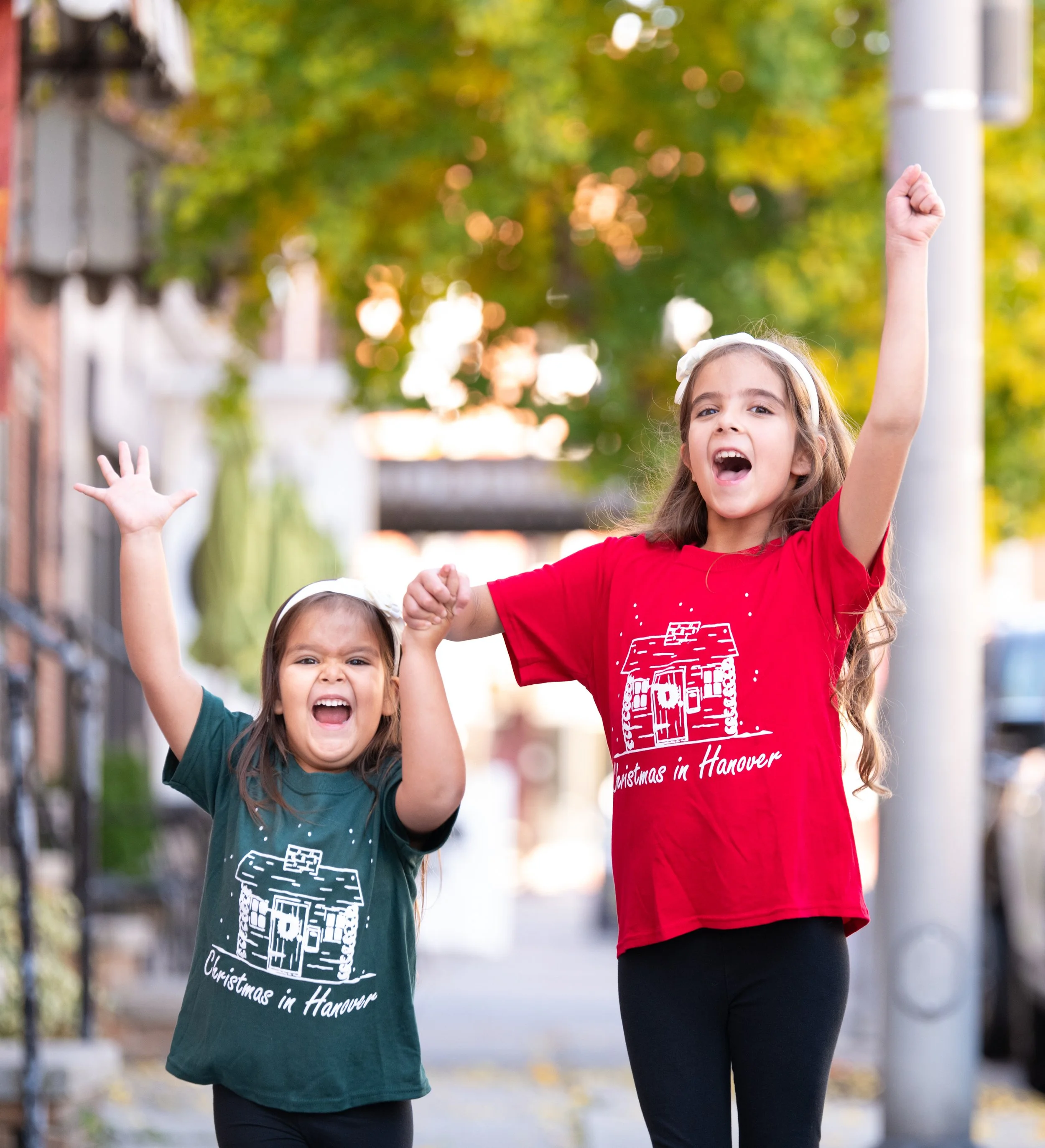 Two young girls walking outdoors, raising their fists in celebration. One girl is wearing a green T-shirt and the other a red T-shirt with a Christmas house design. The background shows trees and a blurred street scene.