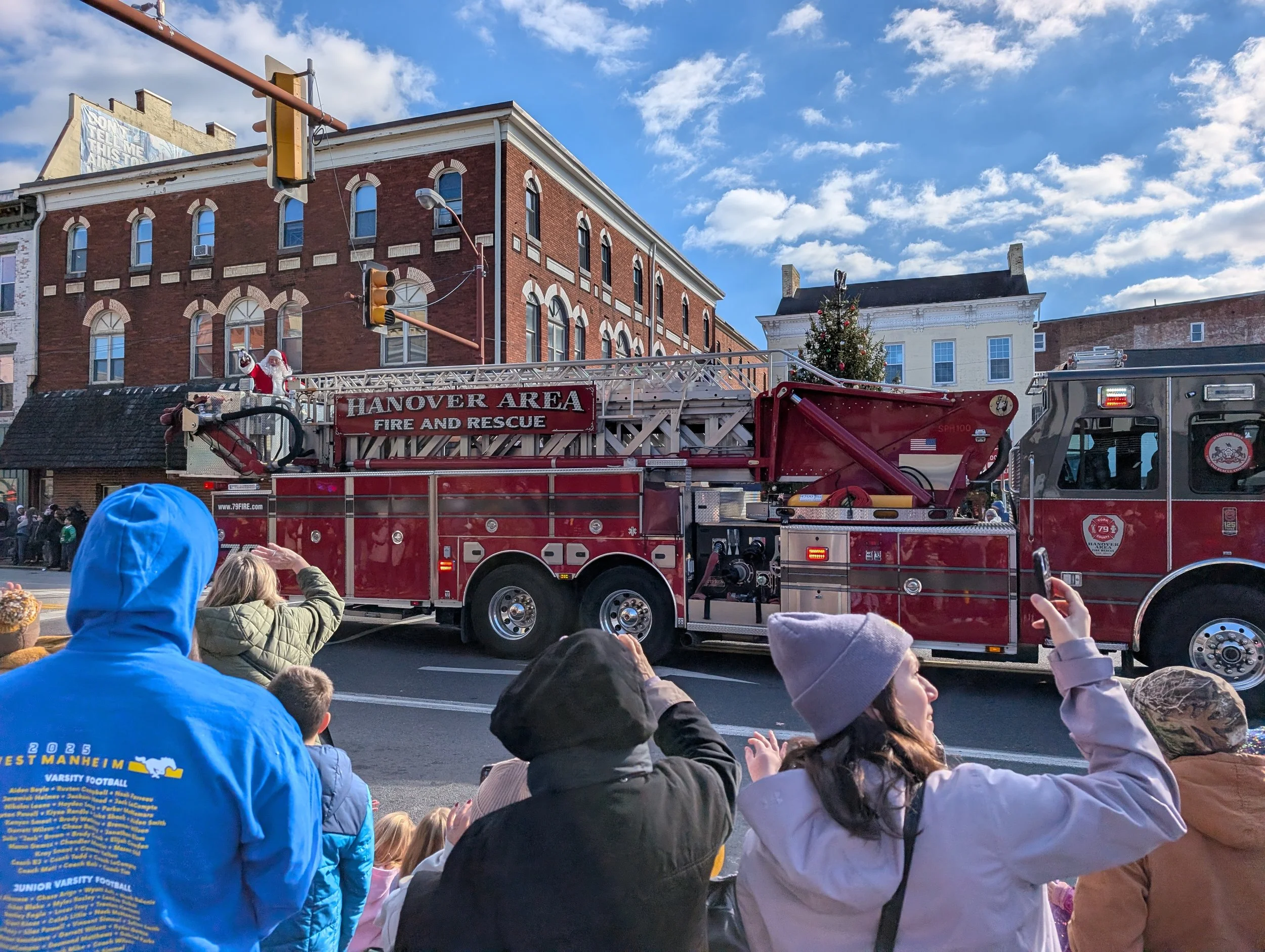 Crowd of people watching a parade with a fire truck that has a Christmas tree on top and a Santa Claus decoration, in front of brick buildings and a blue sky with clouds.