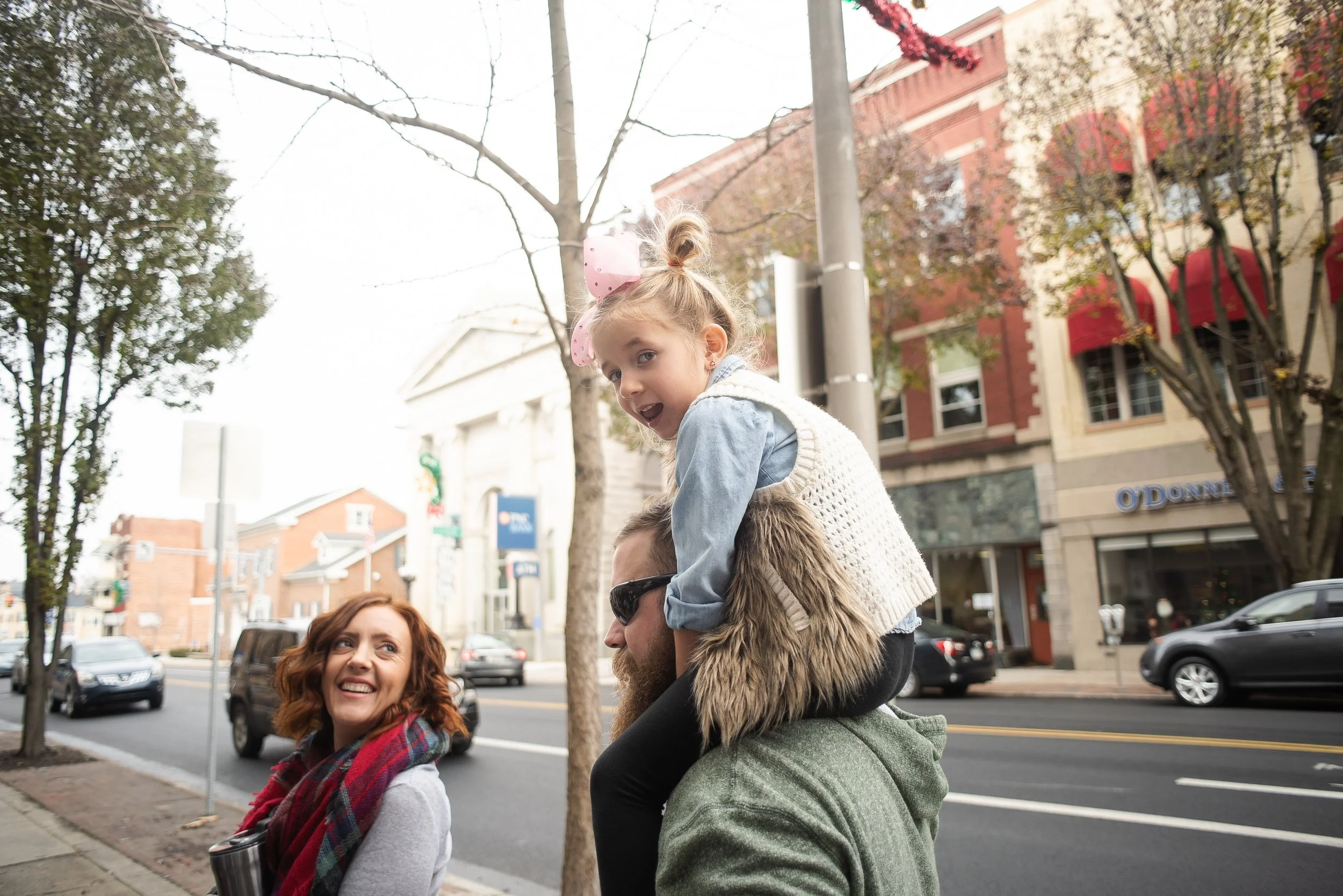 A girl with a pink bow on her hair sitting on a man's shoulders, smiling, while people walk on a city sidewalk with trees and buildings in the background.