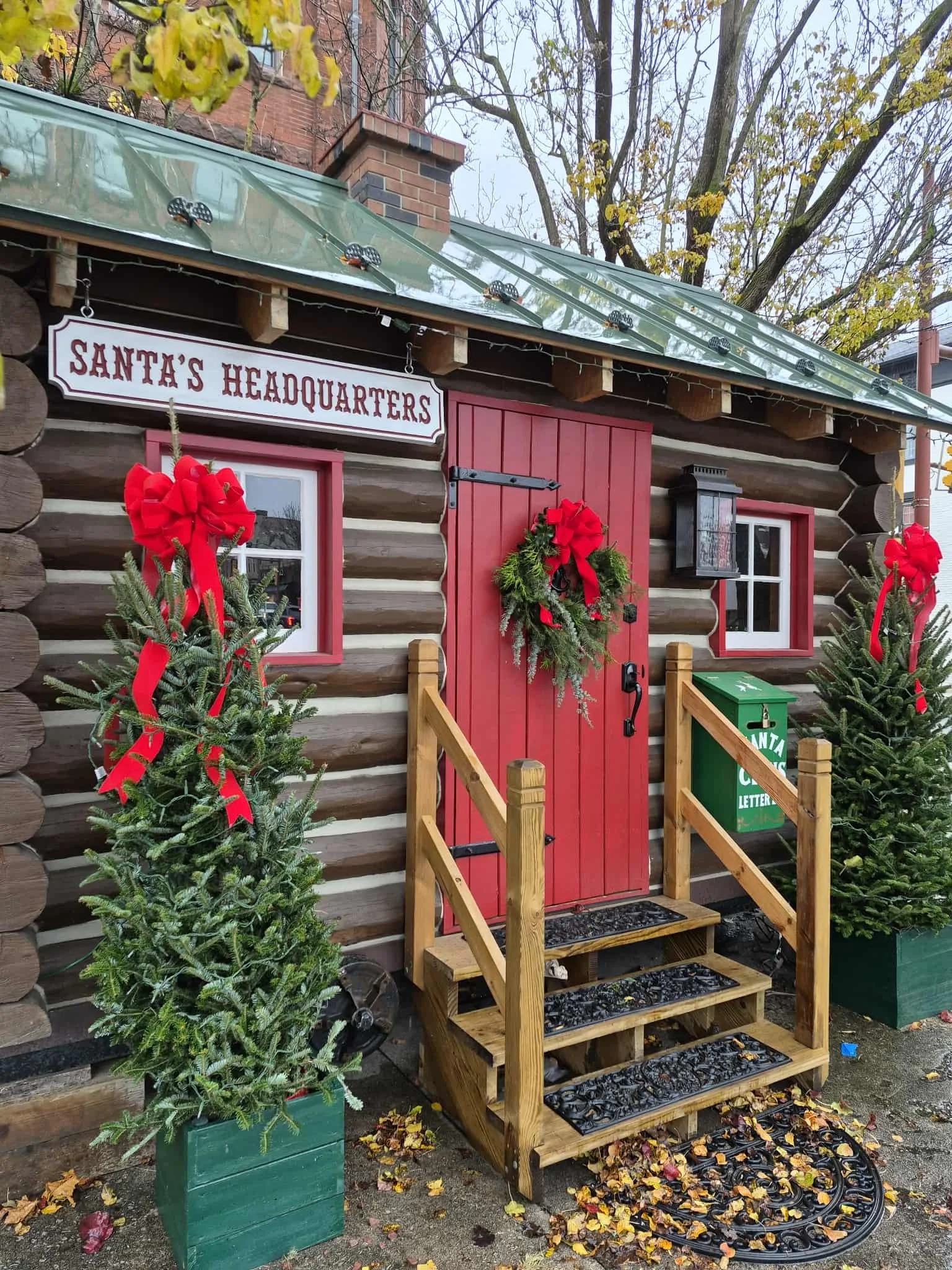 A small wooden building decorated for Christmas with a sign that reads 'Santa's Headquarters,' red door, wreath, and red bows on the windows. Two small Christmas trees with red ribbons and green boxes are on either side of the entrance. The building has a glass roof, and there are trees and fall leaves on the ground.