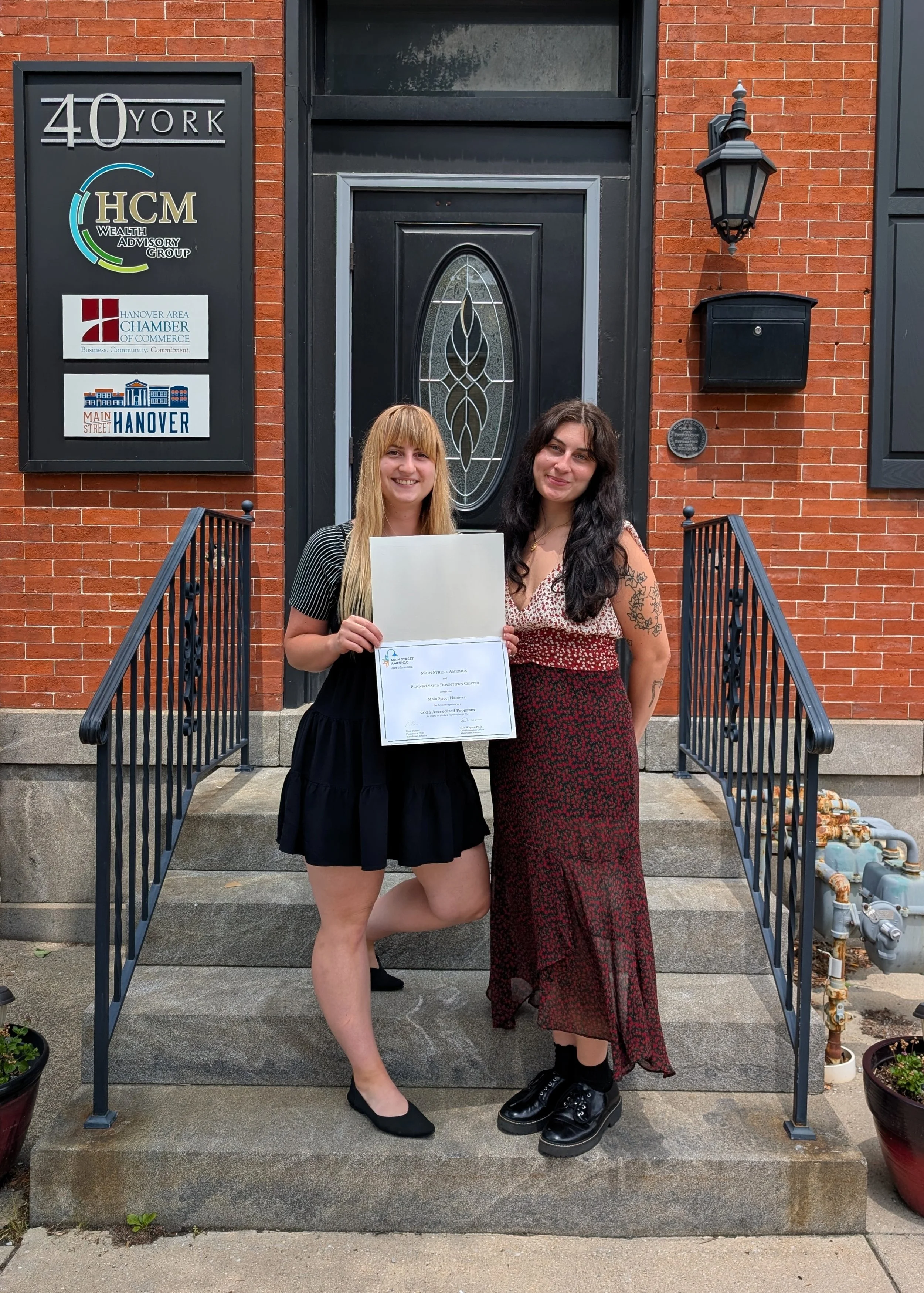 Two women standing on front steps of a brick building, one holding a certificate, smiling. Behind them are signs for local organizations, a black door with a decorative glass panel, a black mailbox, an outdoor lantern, and potted plants.
