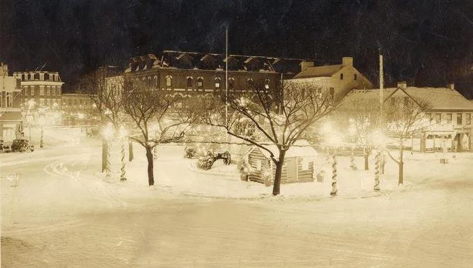 A snowy city street at night with snow-covered ground, leafless trees, parked cars, and illuminated buildings in the background.