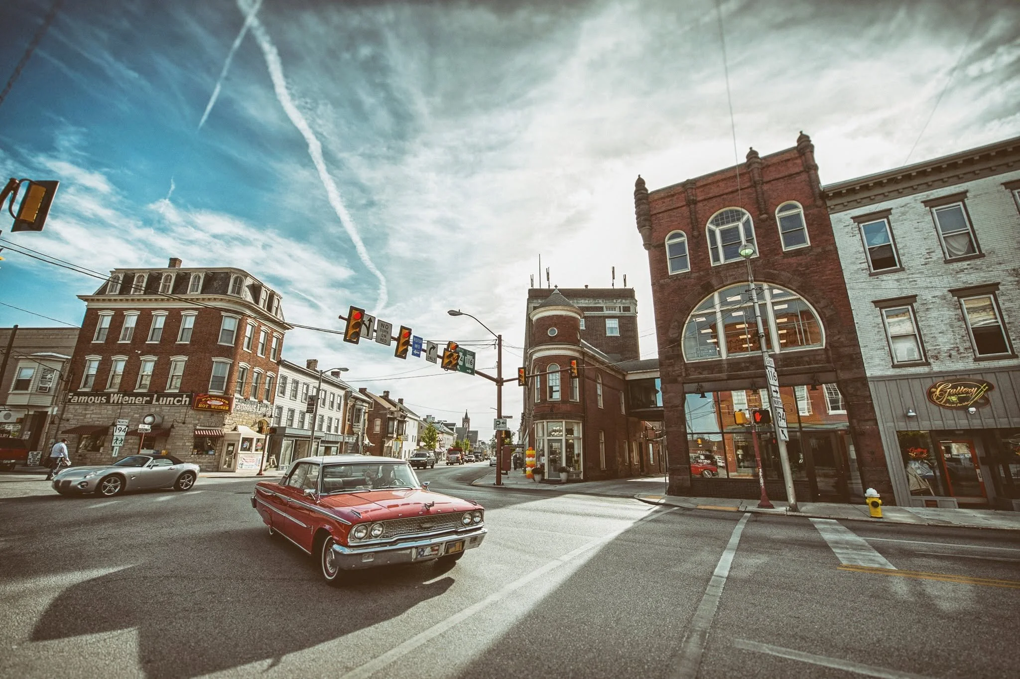 A busy city street with vintage cars parked and driving, historic brick buildings, traffic lights, and storefronts under a partly cloudy sky.