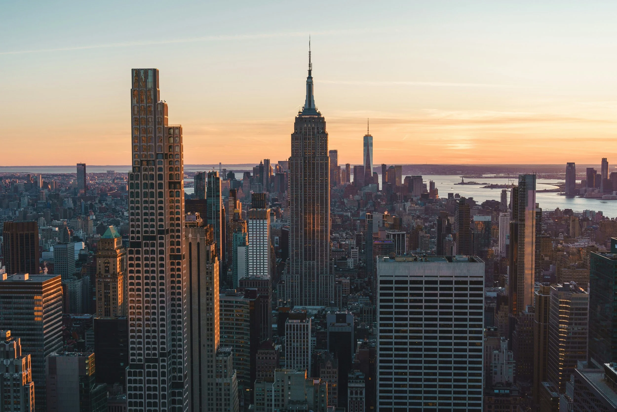 A view of New York City skyline at sunset, featuring the Empire State Building in the center and other skyscrapers, with the Hudson River in the background.
