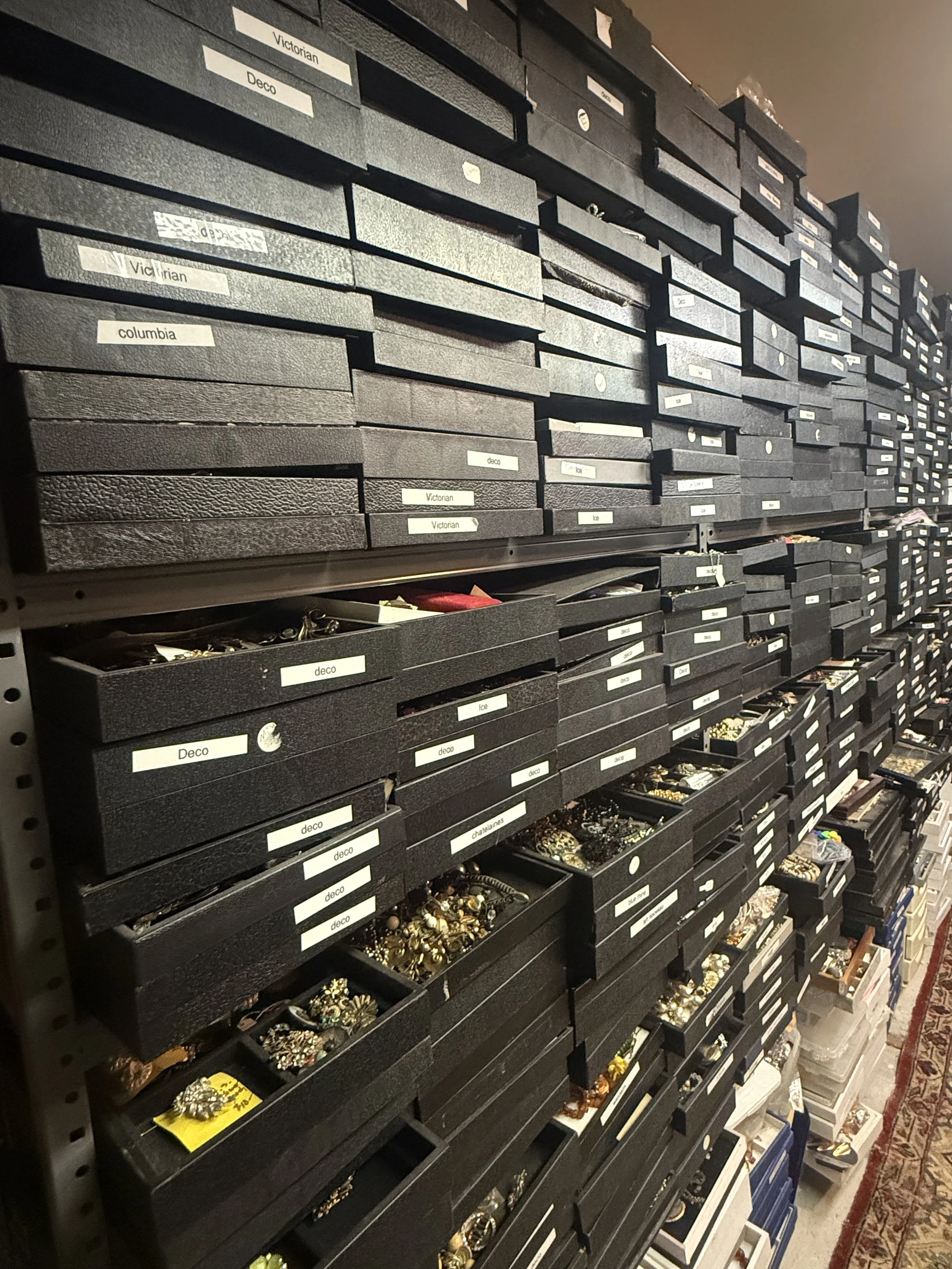 A large shelving unit filled with black storage boxes labeled with words like 'Victorian,' 'Deco,' 'columbia,' and 'ice,' containing various jewelry items, including rings, necklaces, and brooches, in a thrift store or warehouse setting.
