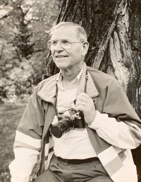 Black and white photograph of artist Rod Stemo pictured leaning against a tree with a camera around his neck.