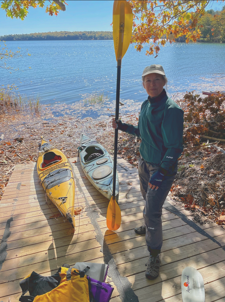 Woman standing next to two kayaks holding an oar in front of a lake
