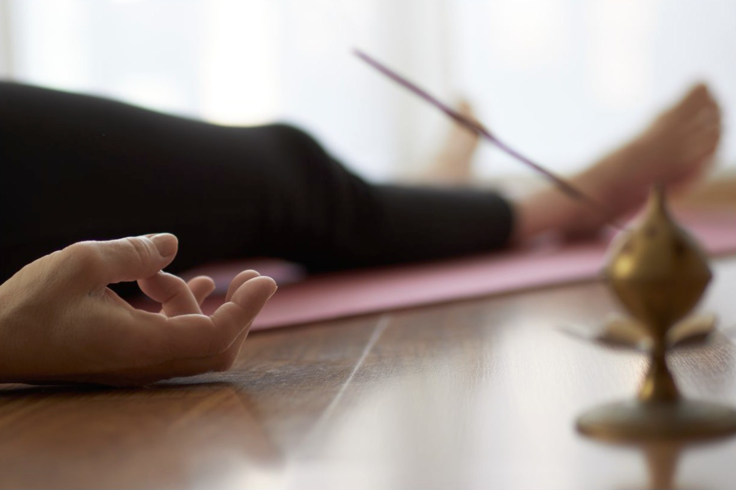 A person practicing meditation with their fingers in a mudra position, lying on a pink yoga mat, with a brass incense holder in the foreground.