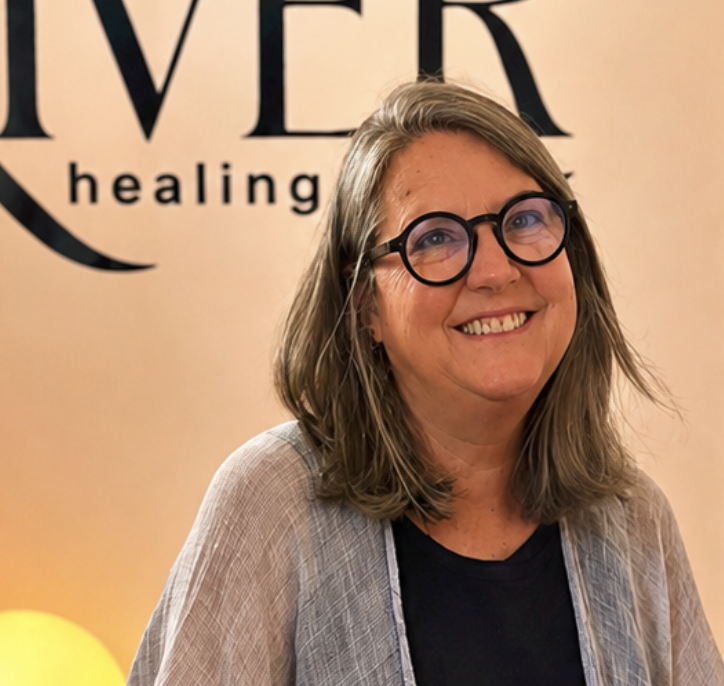 Smiling woman with shoulder-length hair, wearing glasses and a black shirt, standing in front of a backdrop with the words 'OVER healing' visible.