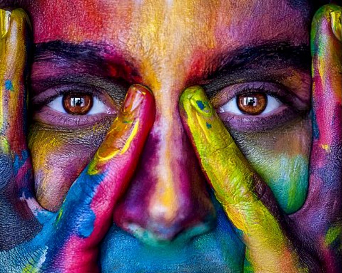 Close-up of a person's face with vibrant paint in multiple colors, holding fingers painted in similar colors over their eyes.