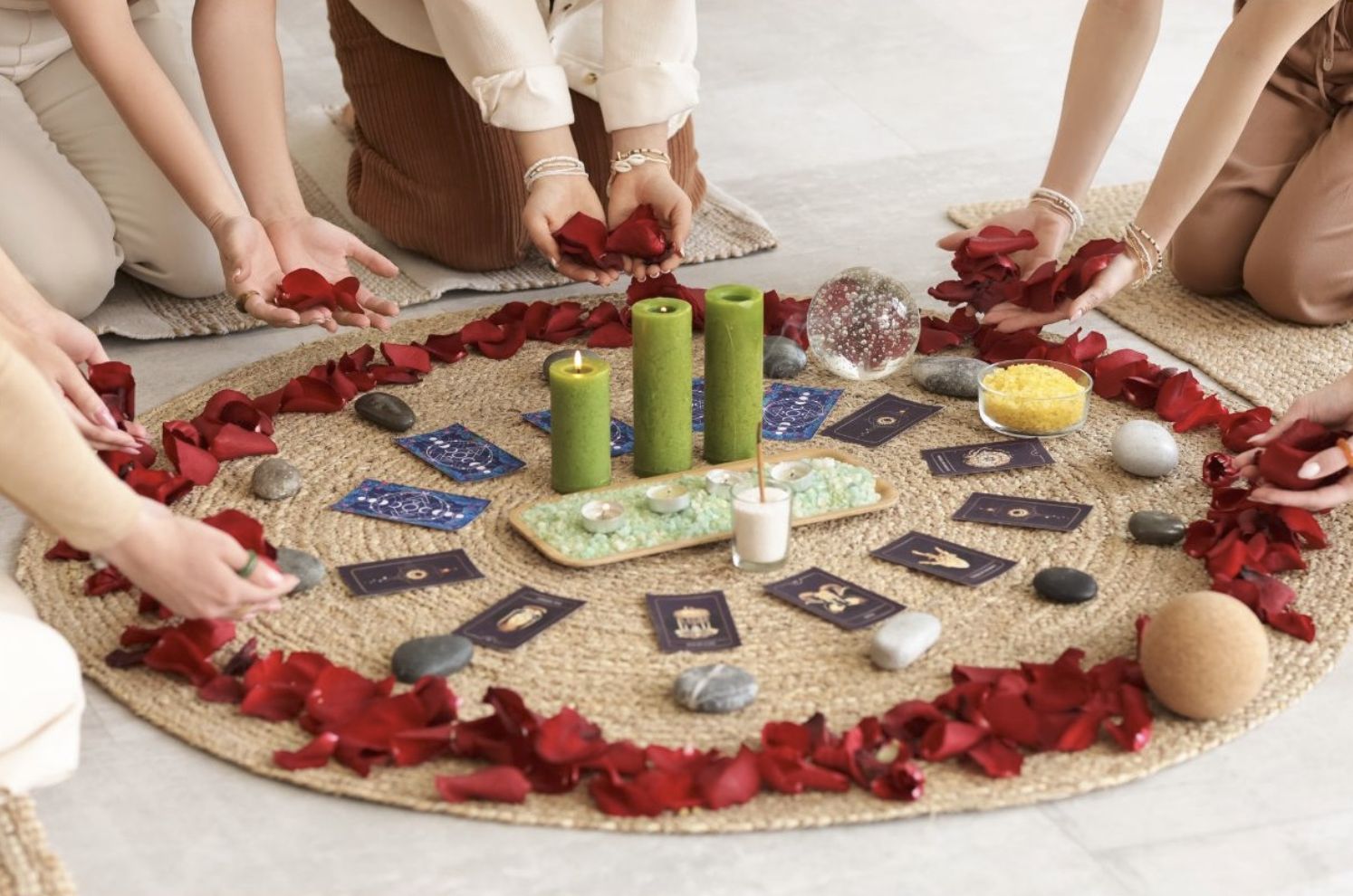 People participating in a spiritual or meditation ritual, seated around a circular arrangement of red rose petals, with candles, tarot cards, crystals, and decorative items in the center.